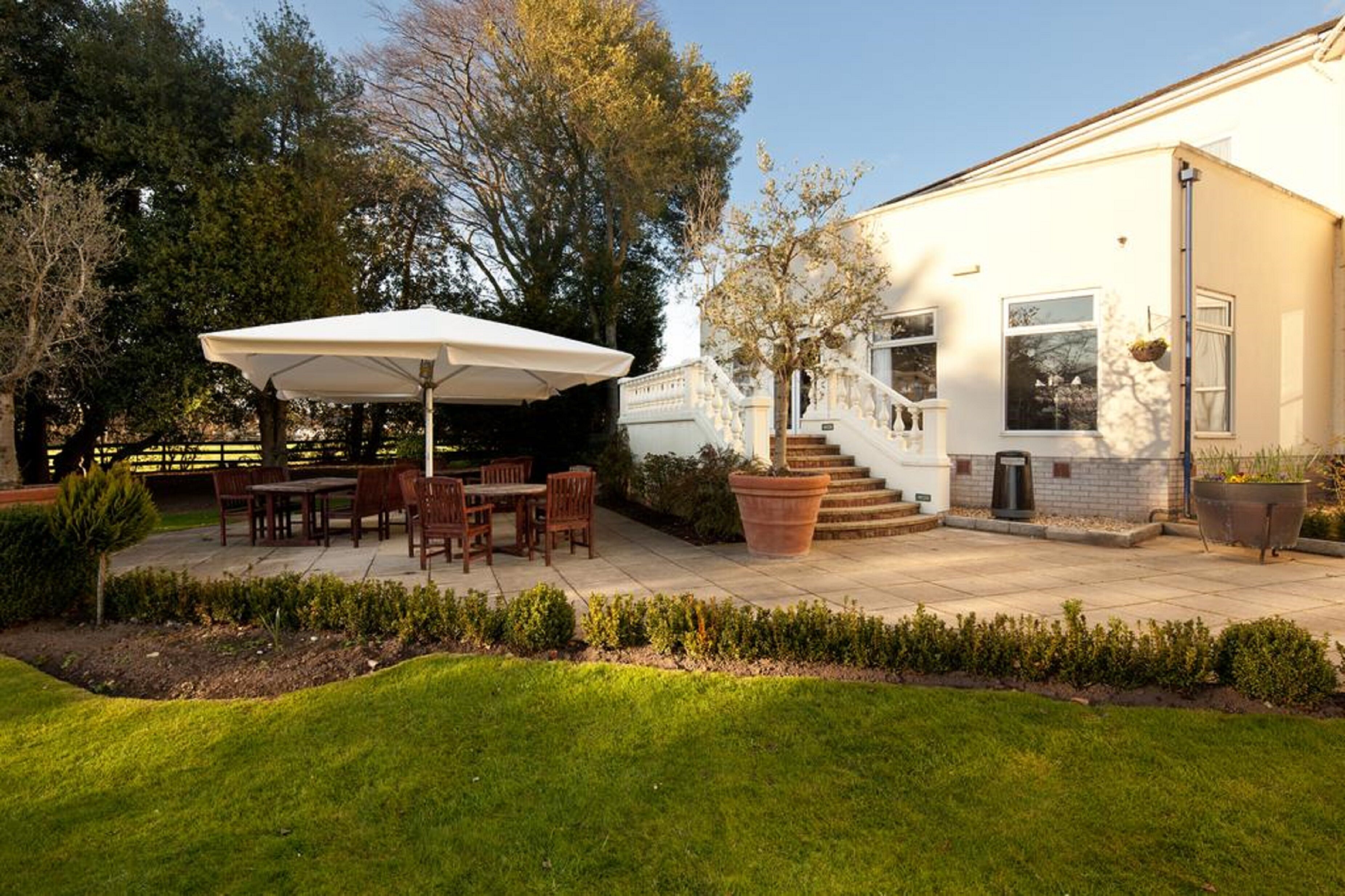 Une terrasse avec des meubles d’extérieur et un grand parasol, une maison blanche avec un escalier et un jardin bien entretenu.