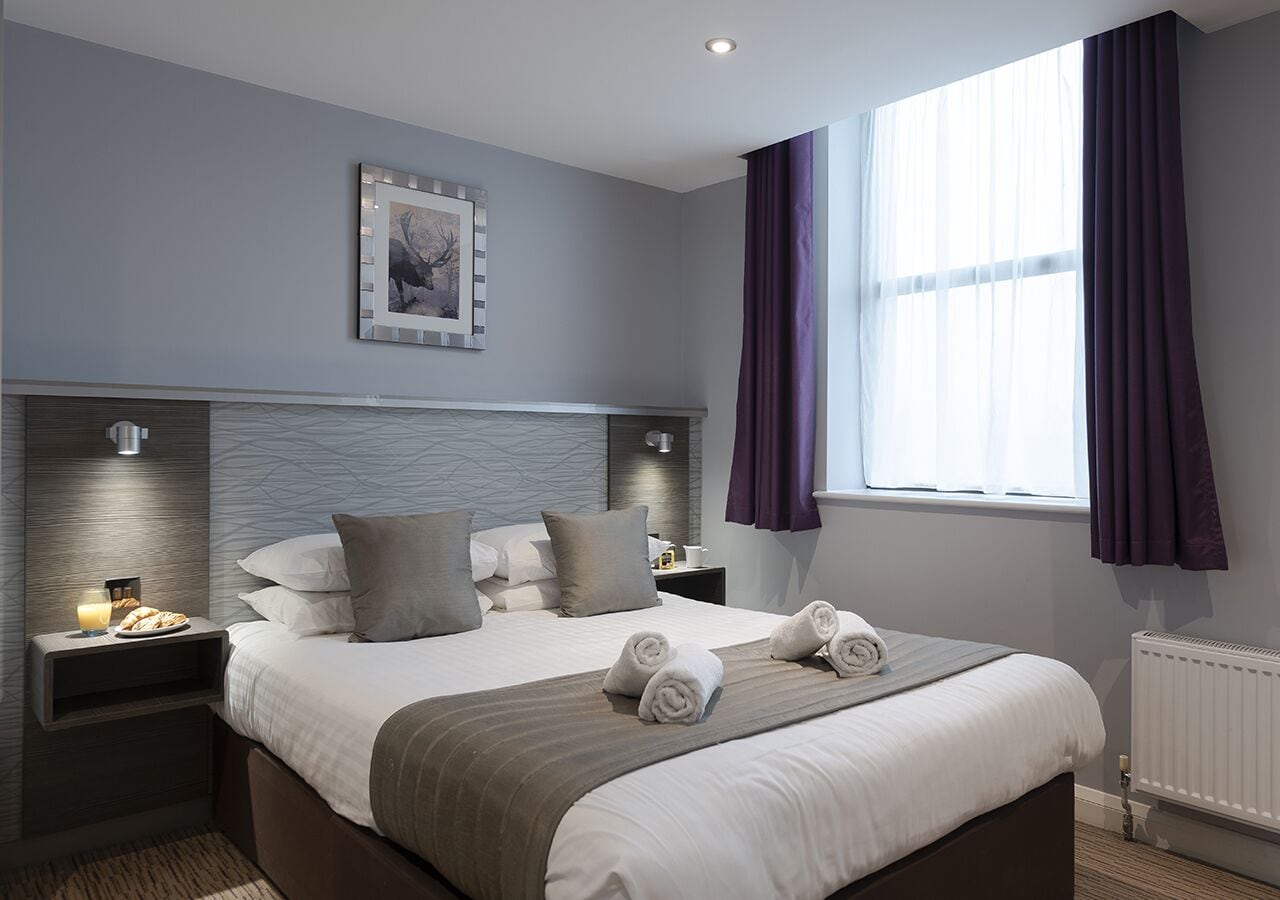 Una habitación de hotel moderna con una cama grande, una mesita de noche con lámpara, un cuadro enmarcado en la pared y una ventana con cortinas.