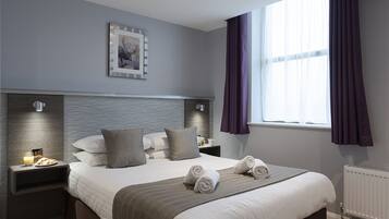 Una habitación de hotel moderna con una cama grande, una mesita de noche con lámpara, un cuadro enmarcado en la pared y una ventana con cortinas.