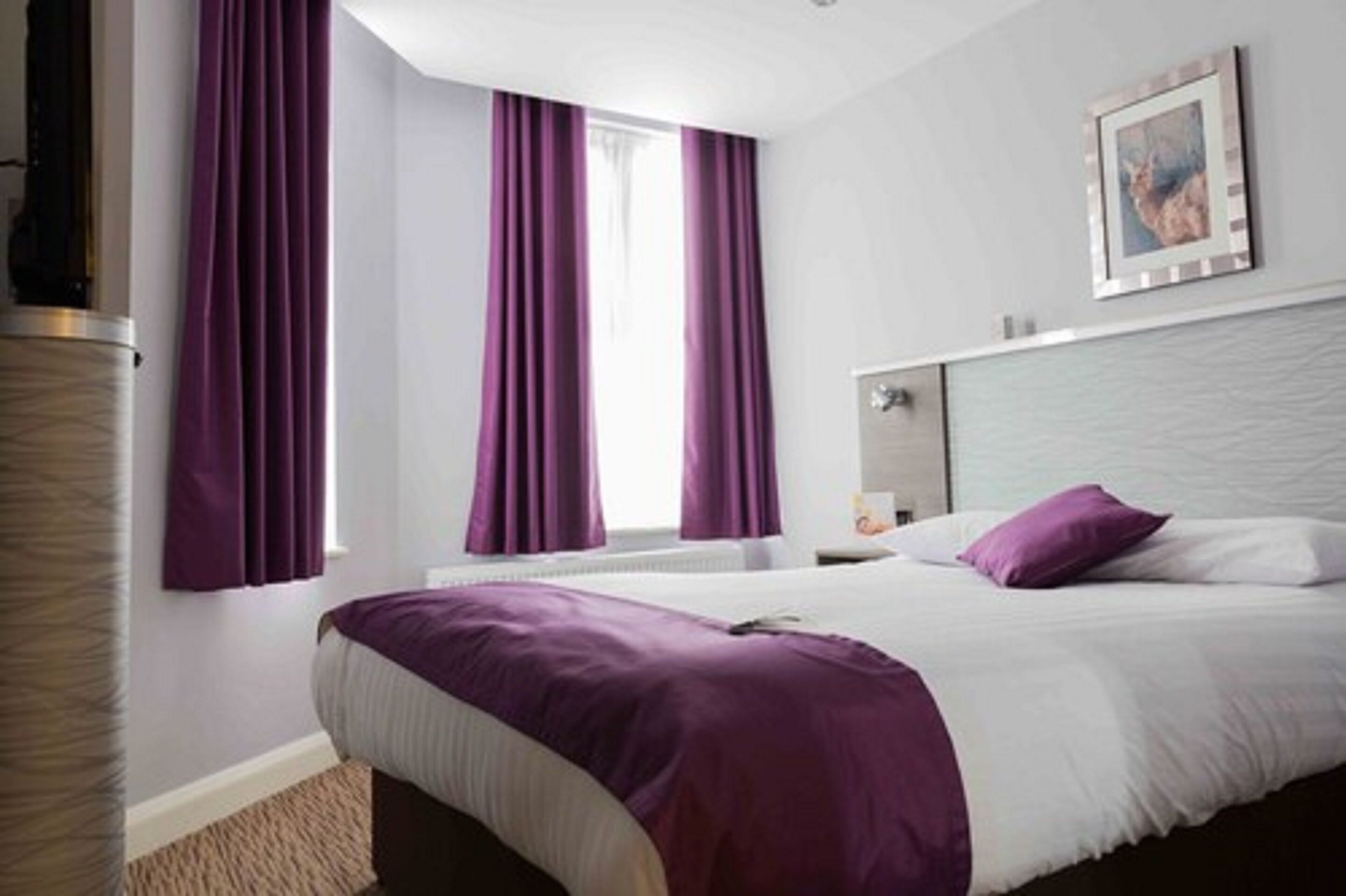 Una camera d'albergo con un letto, tende viola, un quadro appeso e una testiera.