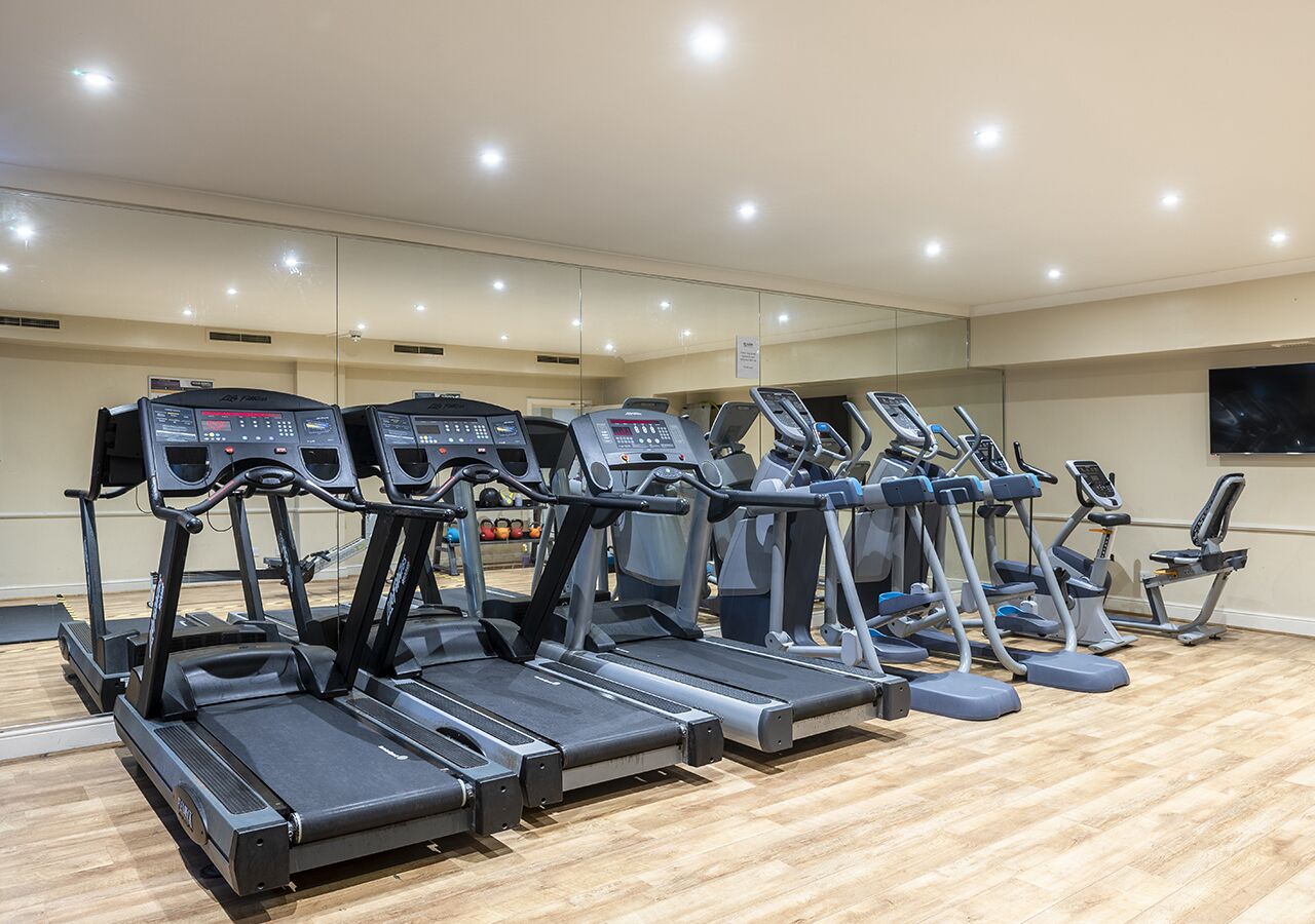 Una hilera de cintas de correr en un gimnasio con suelo de madera y paredes espejadas.