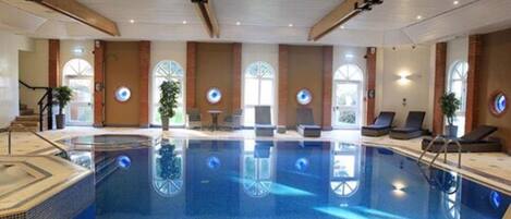 An indoor swimming pool with a tiled floor, a lounge area with chairs, and a staircase leading to another level.