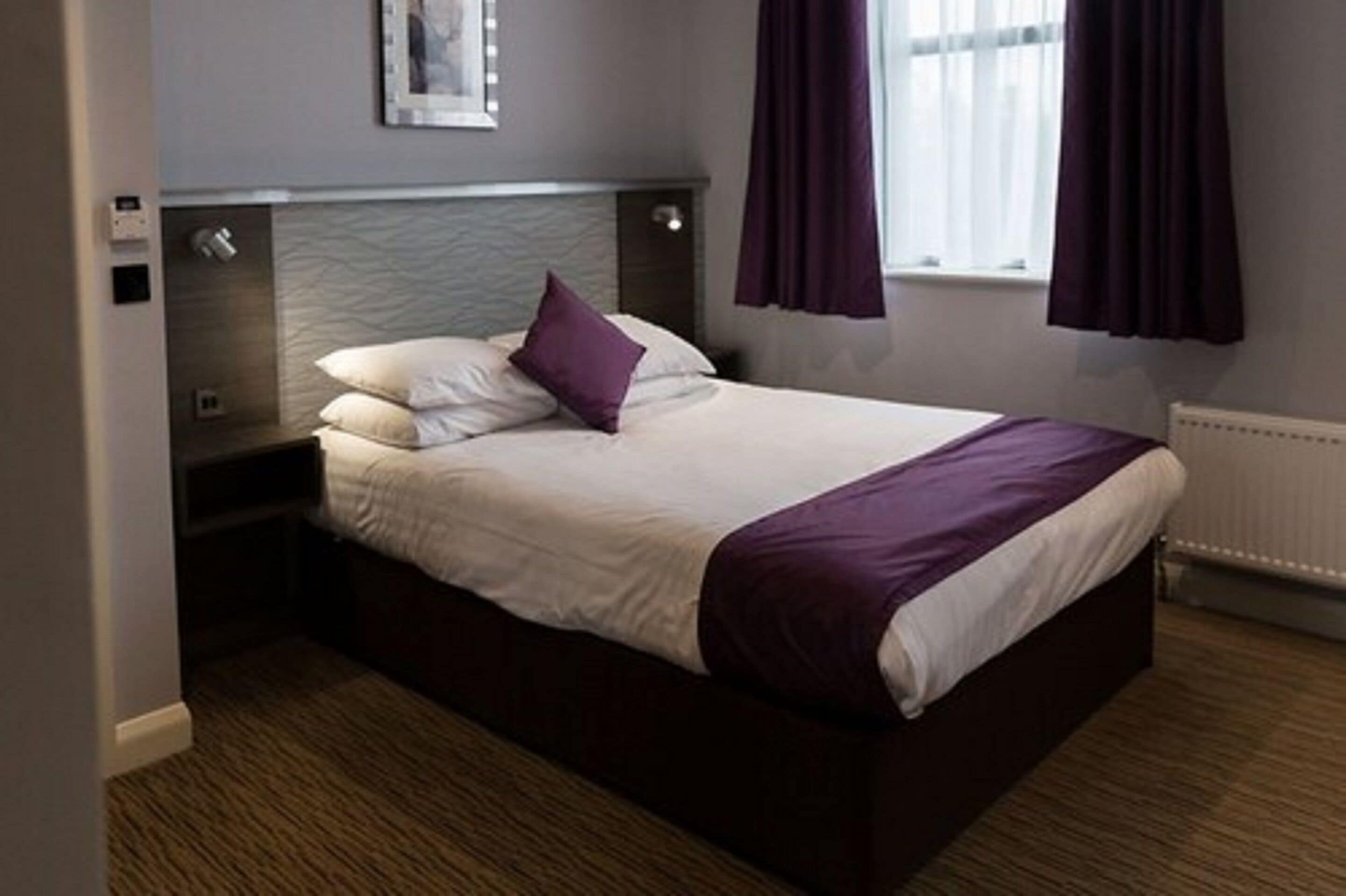 A neatly made bed with a white and purple bedspread, a headboard with a textured design, and a window with purple curtains.