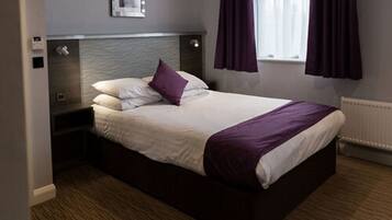 A neatly made bed with a white and purple bedspread, a headboard with a textured design, and a window with purple curtains.