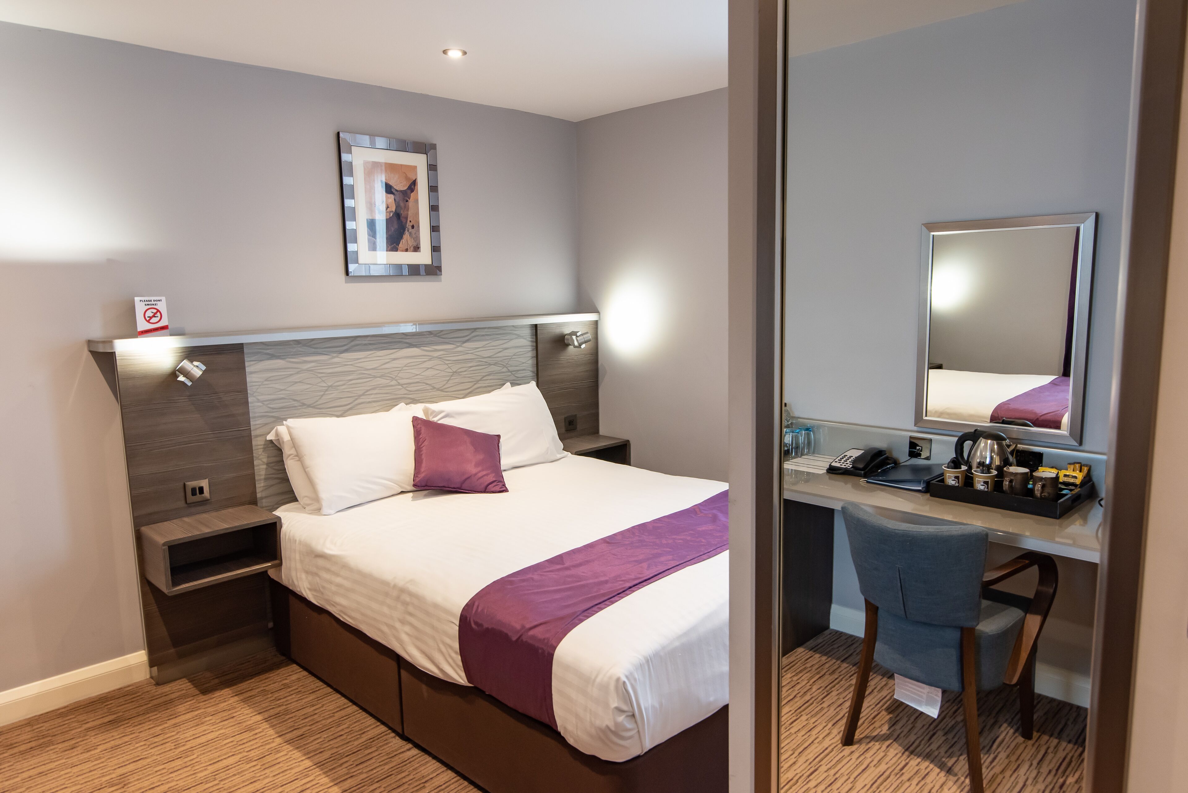 Una habitación de hotel moderna con una cama bien hecha, un escritorio con silla, una mesita de noche y un espejo grande.