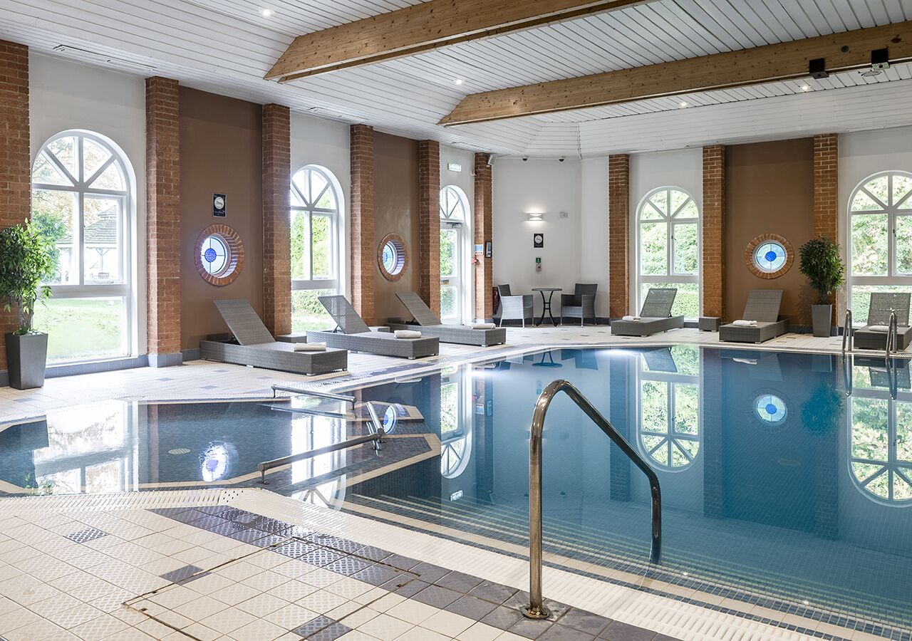 An indoor swimming pool with clear glass walls, lounge chairs, and a sunlit room with large windows.