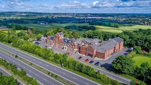 An aerial view of a large, historic building with a prominent red brick facade, surrounded by lush greenery and a busy road. - The Hog's Back Hotel & Spa Farnham (Farnham)
