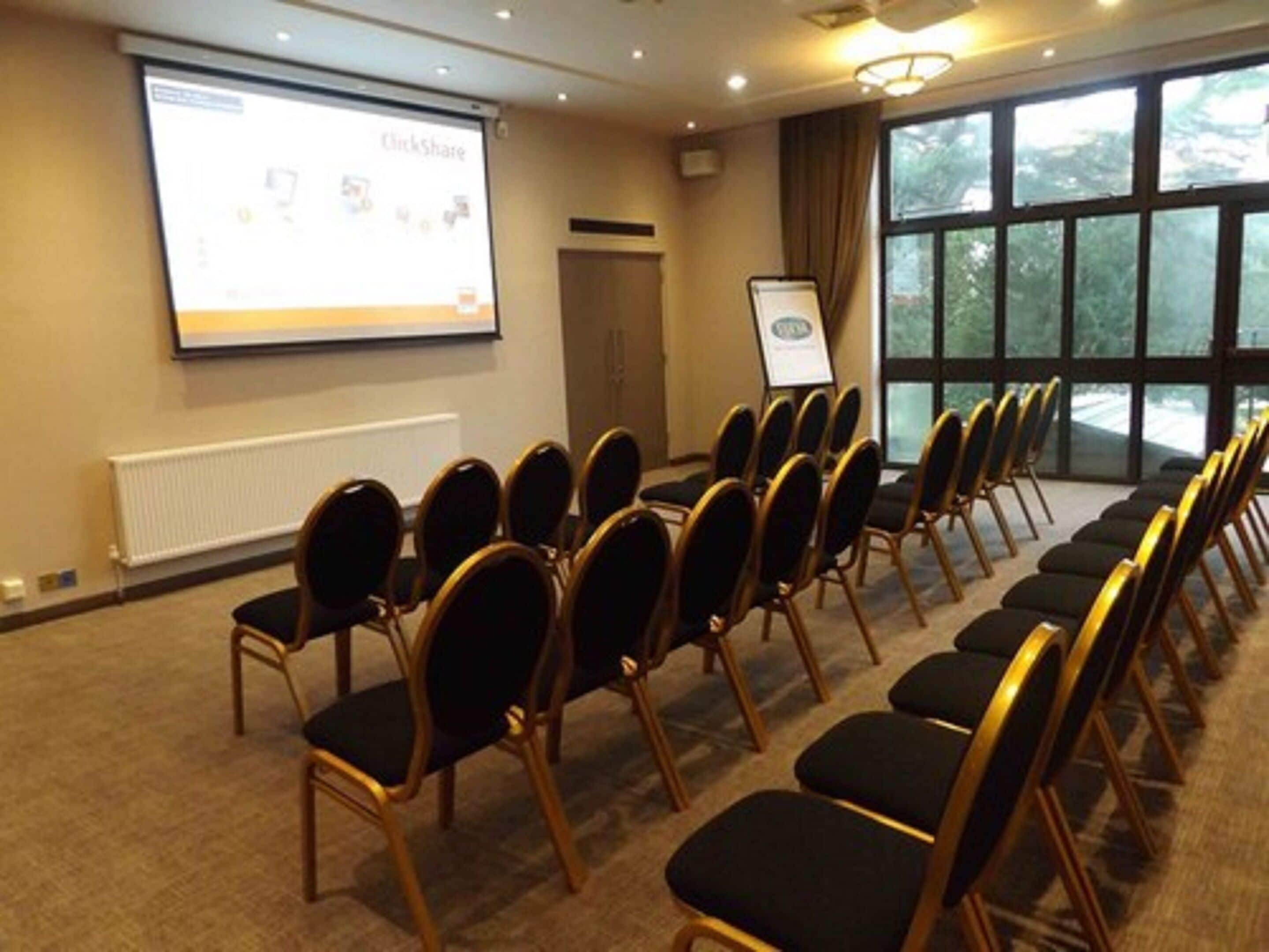 A conference room with rows of chairs facing a projection screen, a whiteboard, and a window with a view of trees.