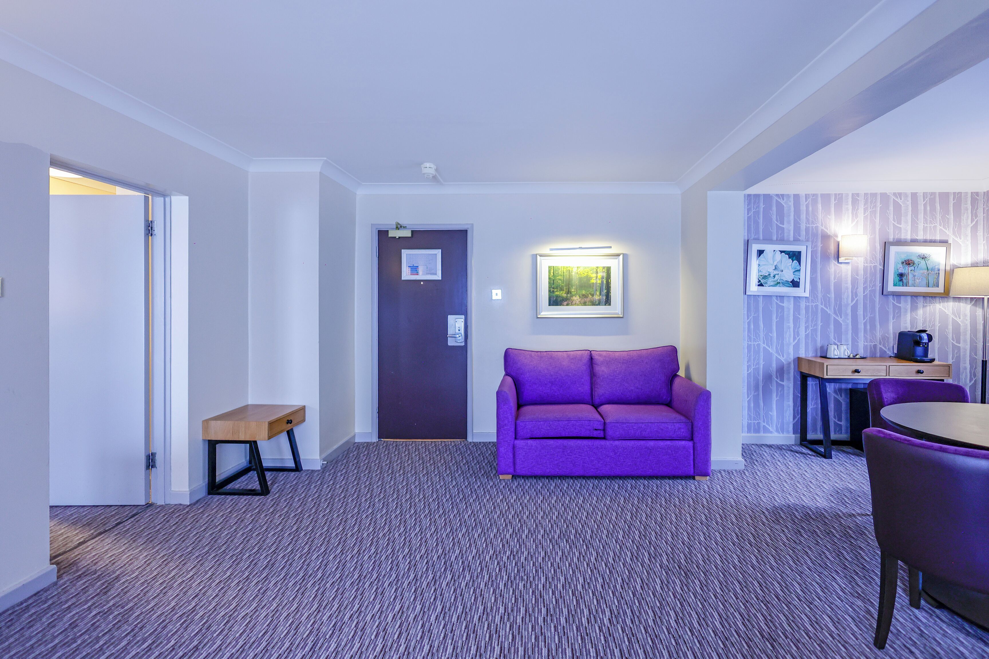 A hotel room with a purple sofa, a small wooden bench, a desk with a chair, and a wall-mounted lamp.