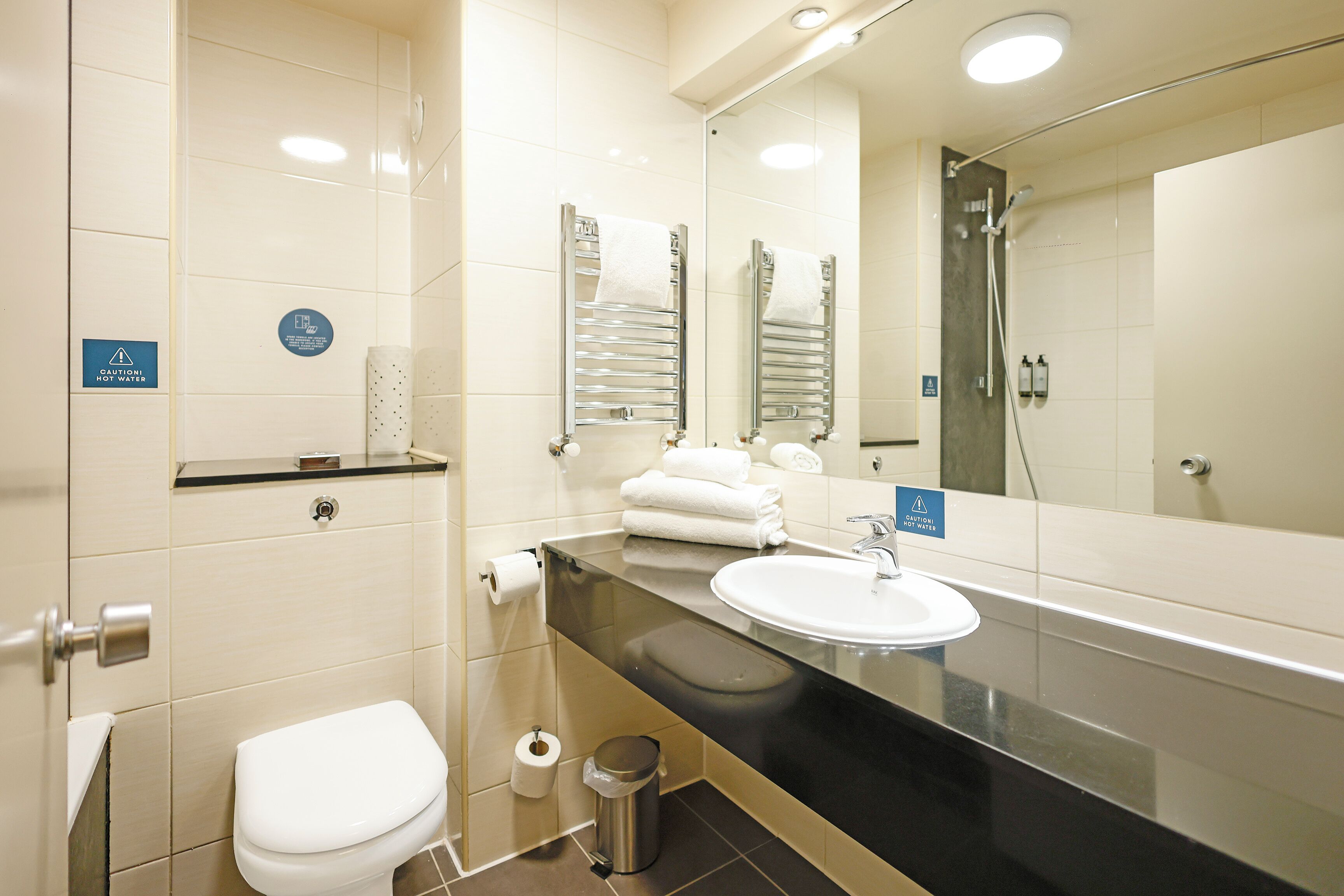 A modern bathroom with a white toilet, a sink, and a mirror. There are towels and a trash can visible.