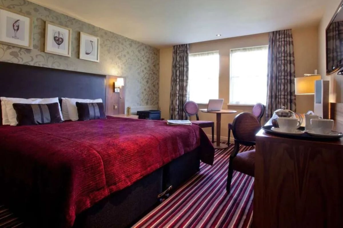 A hotel room with a large bed, a desk with a chair, a laptop, and a table with a teapot and cups.