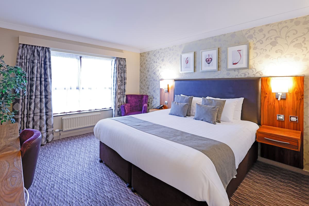A hotel room with a large bed, a nightstand, a chair, and a window with curtains.