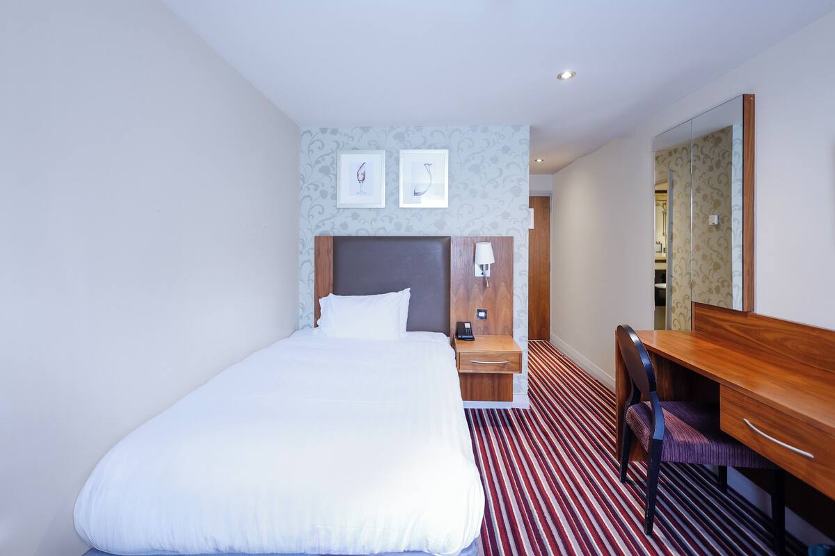 A hotel room with a large bed, a wooden desk, a chair, a nightstand, and a mirror.