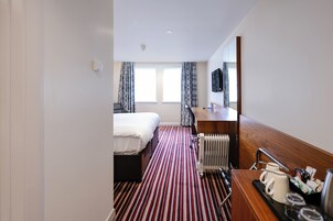 Una habitación de hotel con una cama, un aparador de madera, un radiador y una ventana con cortinas.