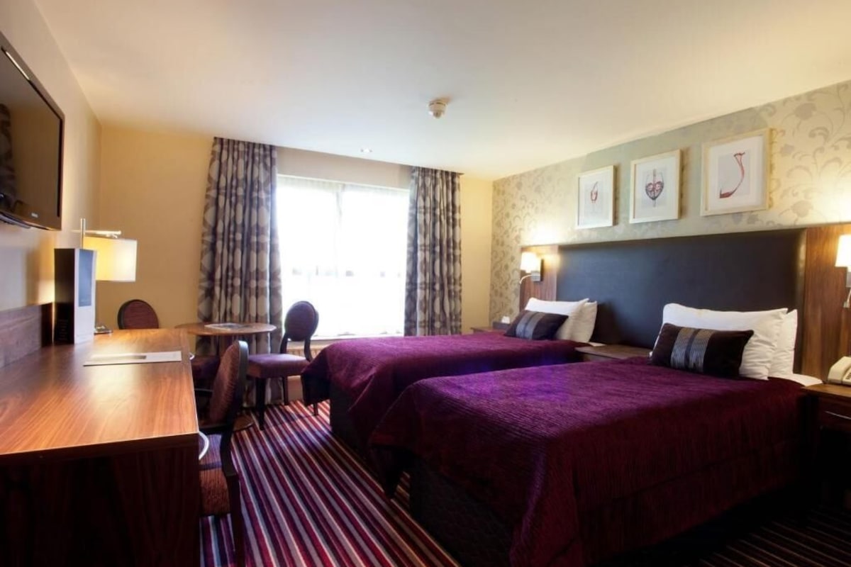 A hotel room with two beds, a desk, a chair, a TV, and a window with curtains.