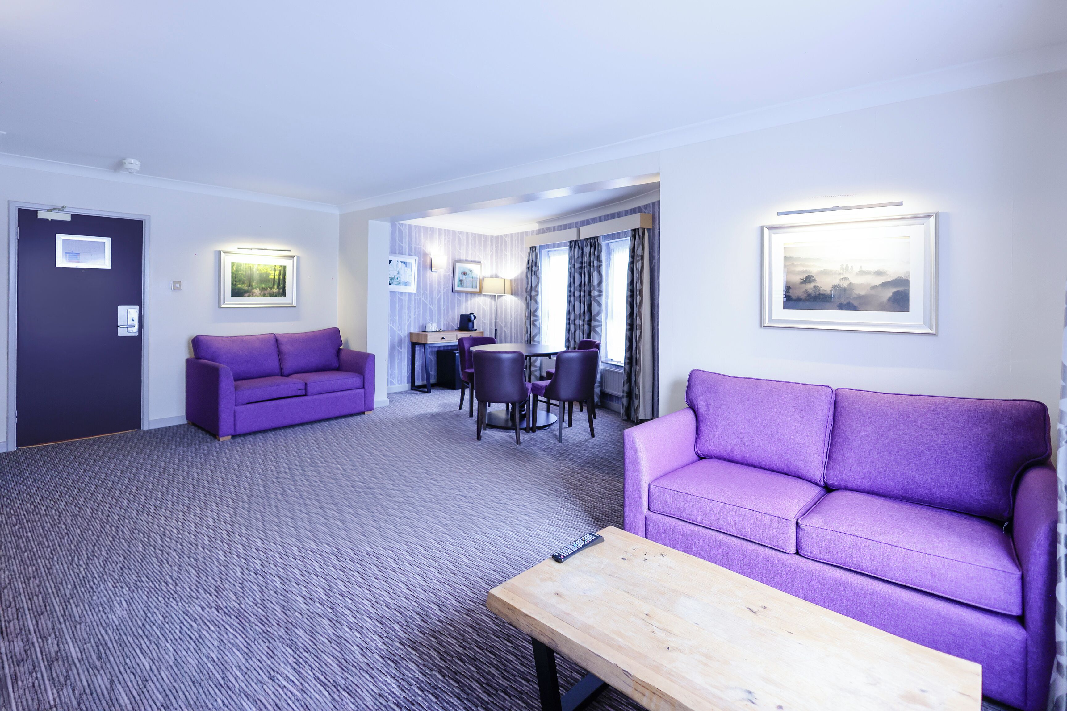 Una habitación de hotel con dos sofás morados, una mesa de centro de madera, una zona de comedor con mesa y sillas, y cuadros enmarcados en las paredes.