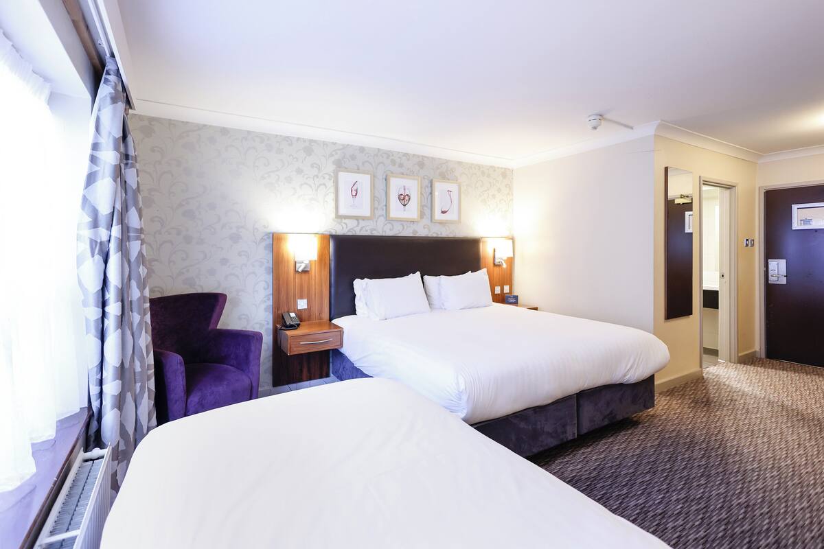 A hotel room with two beds, a chair, a nightstand, and a wall with framed pictures.