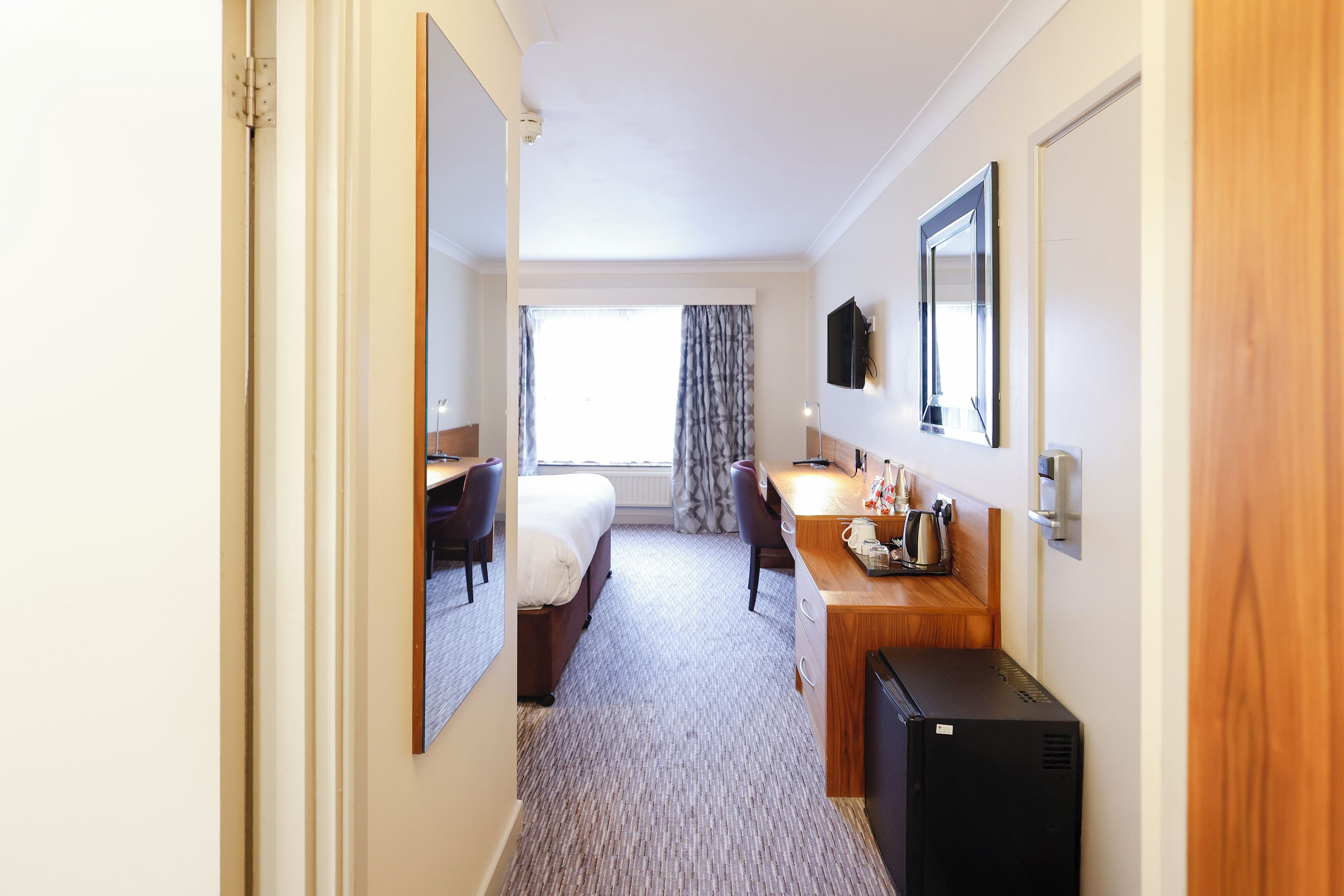 A hotel room with a bed, a desk, a chair, a TV, and a minibar.