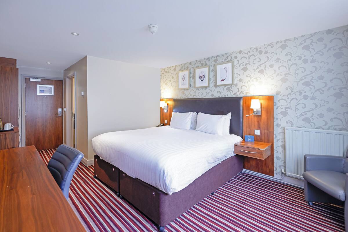 A hotel room with a large bed, a wooden desk, a chair, and a nightstand with a lamp.