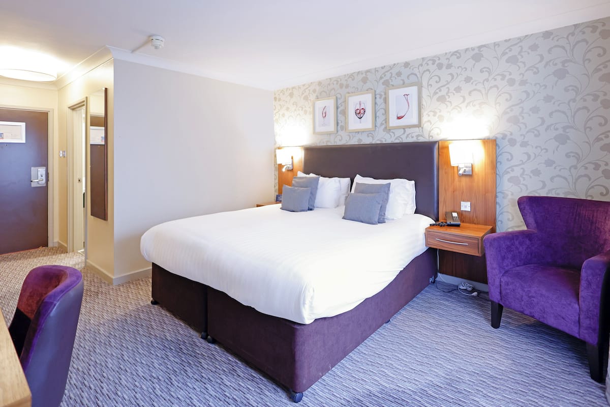 A hotel room with a large bed, a purple armchair, a nightstand with a lamp, and a wall with framed artwork.