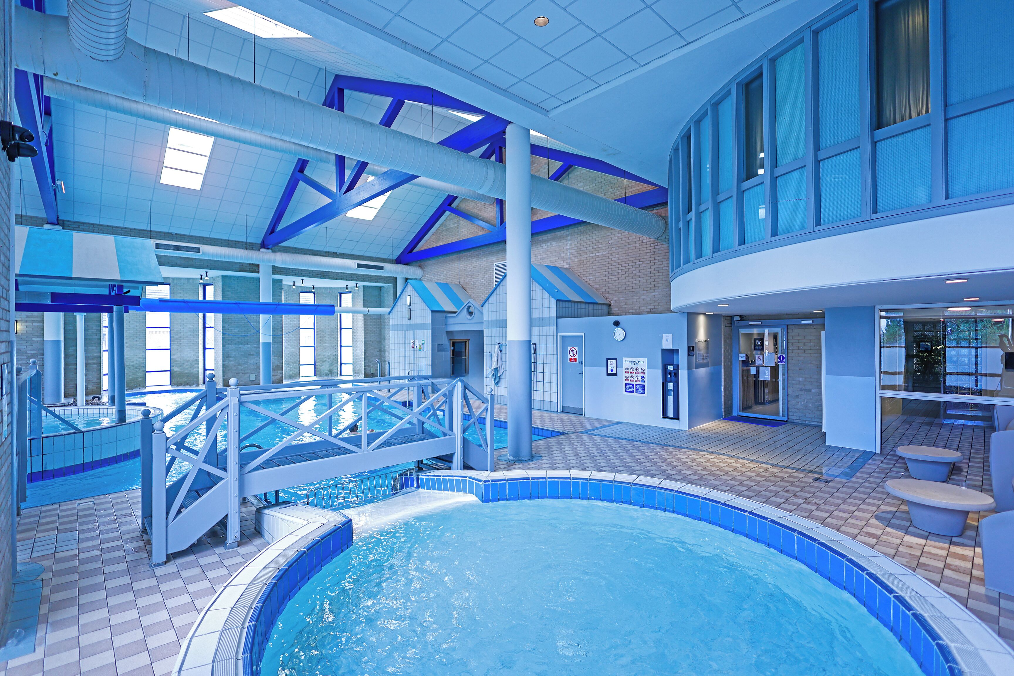 An indoor swimming pool with a blue and white color scheme, a covered bridge, and a seating area with a view of the pool.