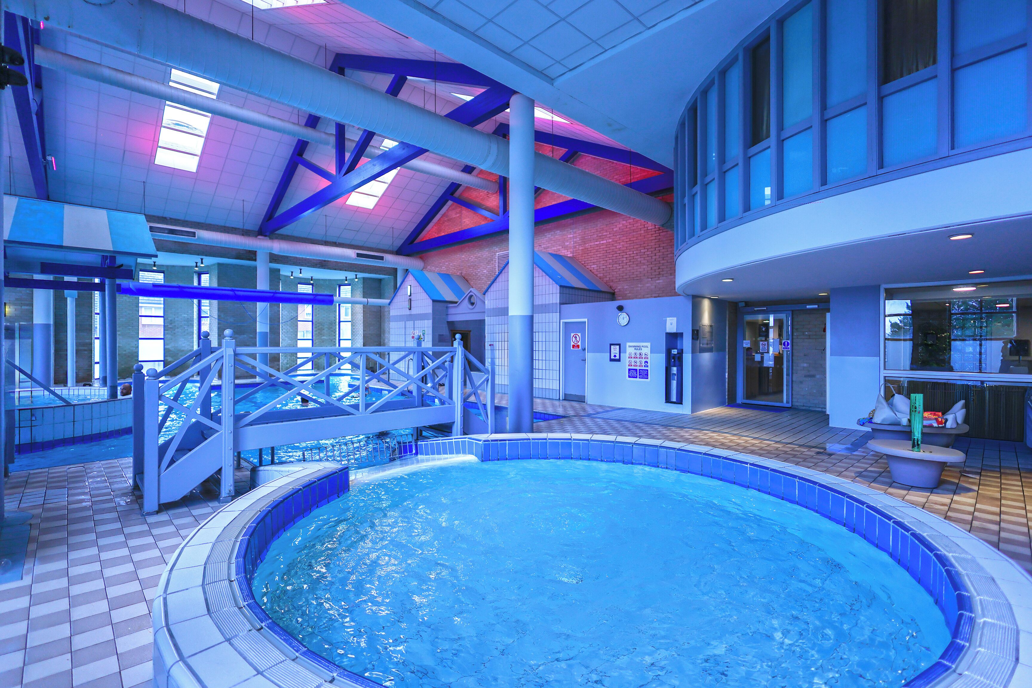 An indoor swimming pool with a tiled floor, a bridge, and a seating area with a view of the pool.