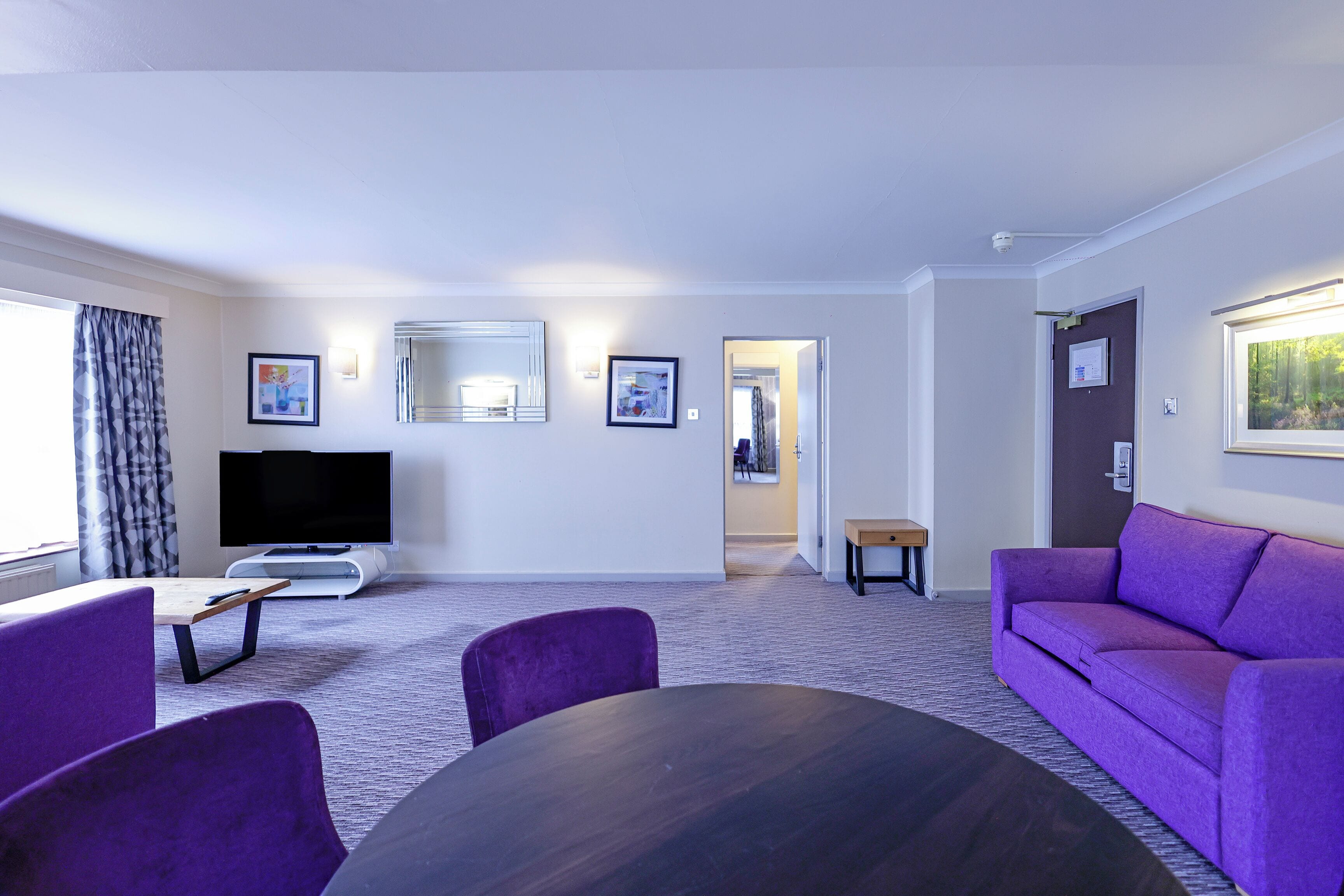 Una sala de estar moderna con un televisor de pantalla plana, muebles morados y un ventanal grande.