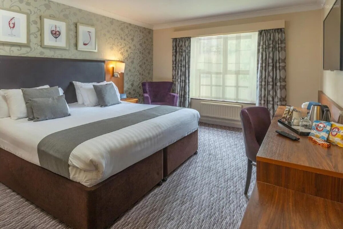 A hotel room with a large bed, a desk with a chair, a window with curtains, and framed artwork on the wall.