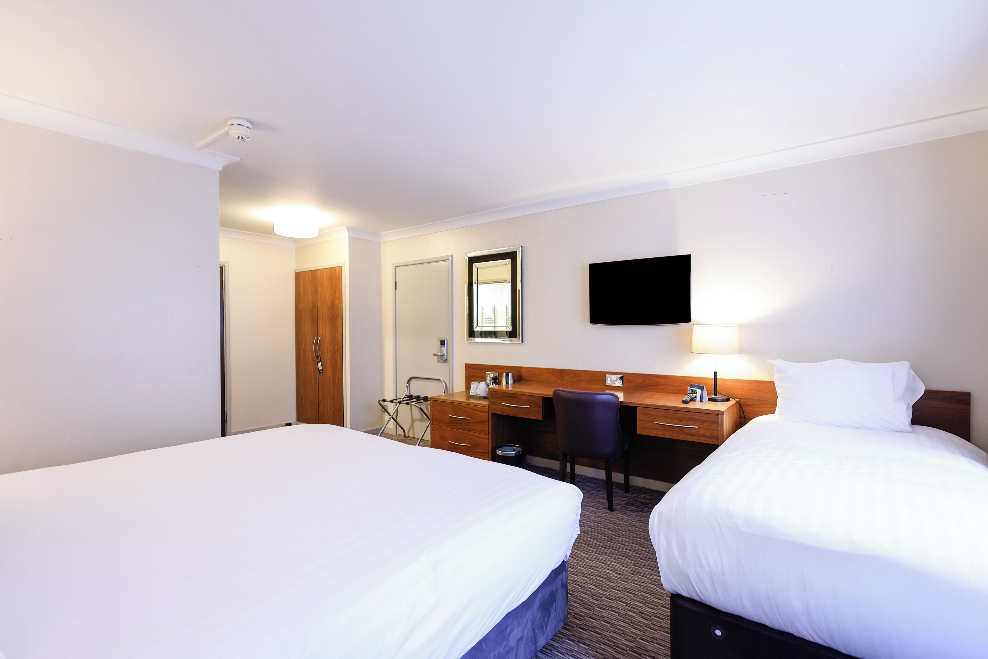 Quarto de hotel com duas camas, uma escrivaninha, uma cadeira, uma televisão e um espelho.