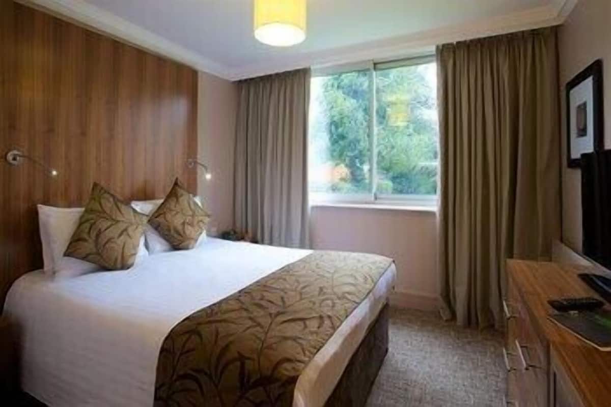 Habitación de hotel con cama, escritorio, ventana con cortinas y televisor montado en la pared.
