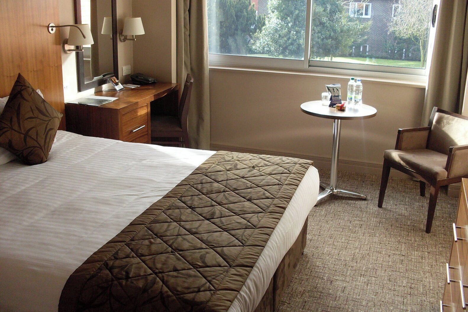Une chambre d’hôtel avec un lit, un bureau, une chaise, une petite table avec des bouteilles d’eau et une fenêtre donnant sur des arbres.