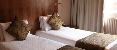 Two single beds with white linens and brown patterned bedspreads, a wooden headboard, and a window with curtains.