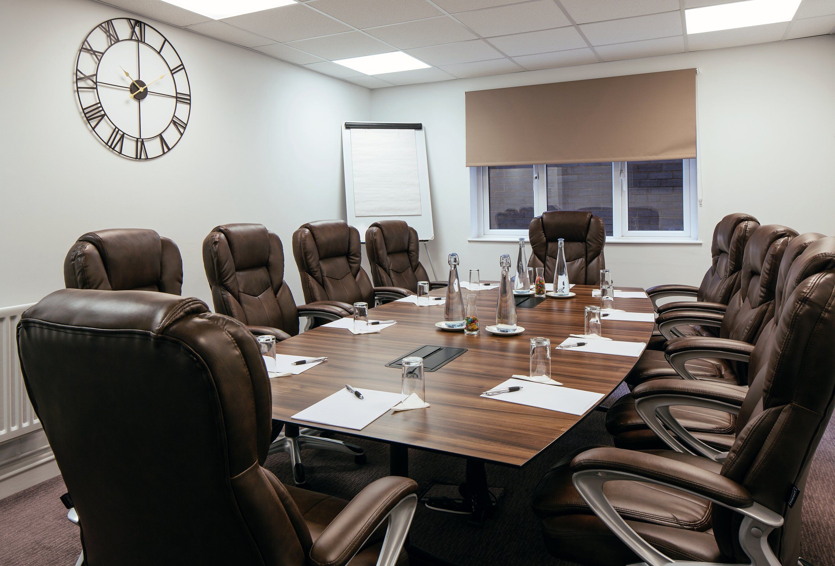 A conference room with a large wooden table, brown leather chairs, a clock, a whiteboard, and a window with blinds.
