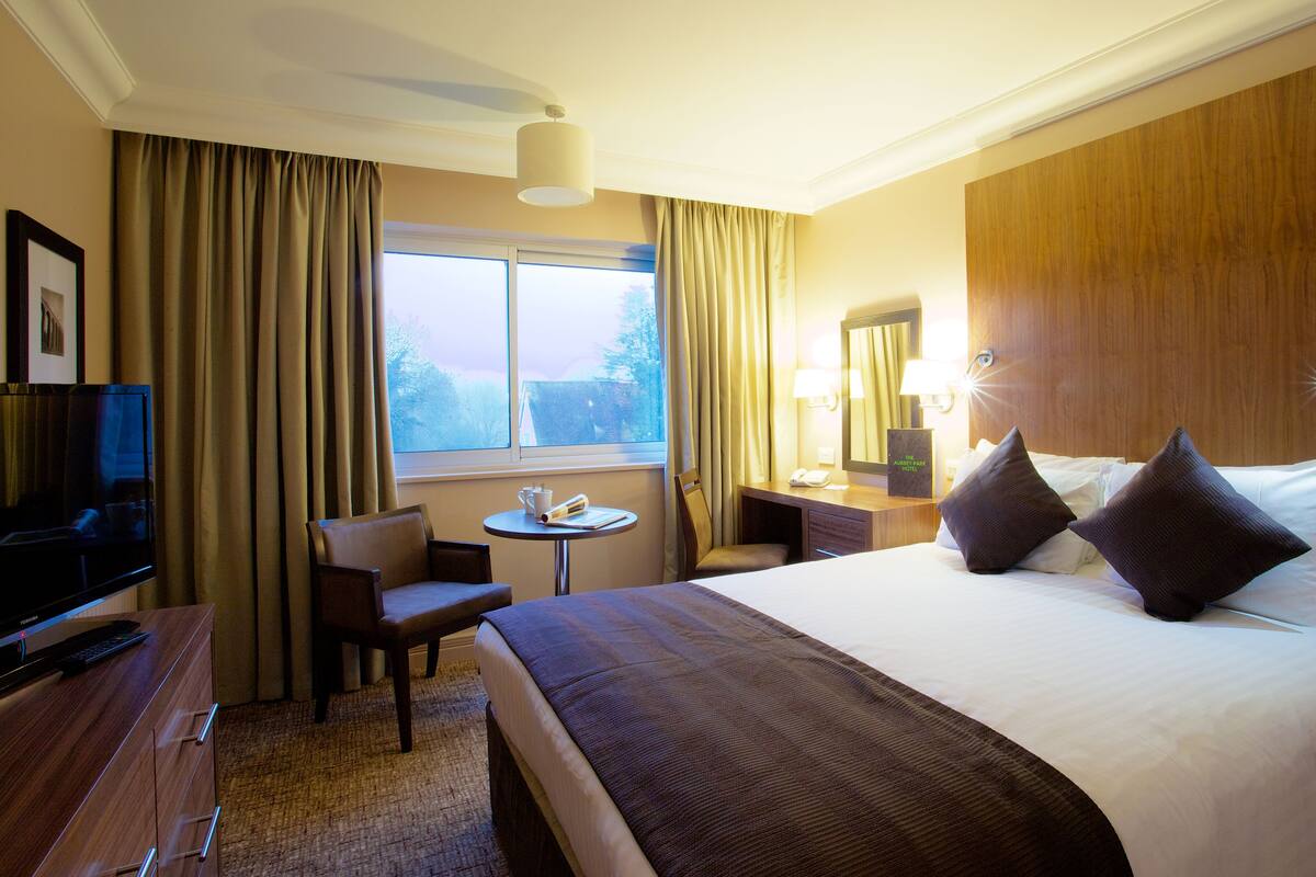 Habitación de hotel con cama, escritorio, silla, televisor y ventana con cortinas.