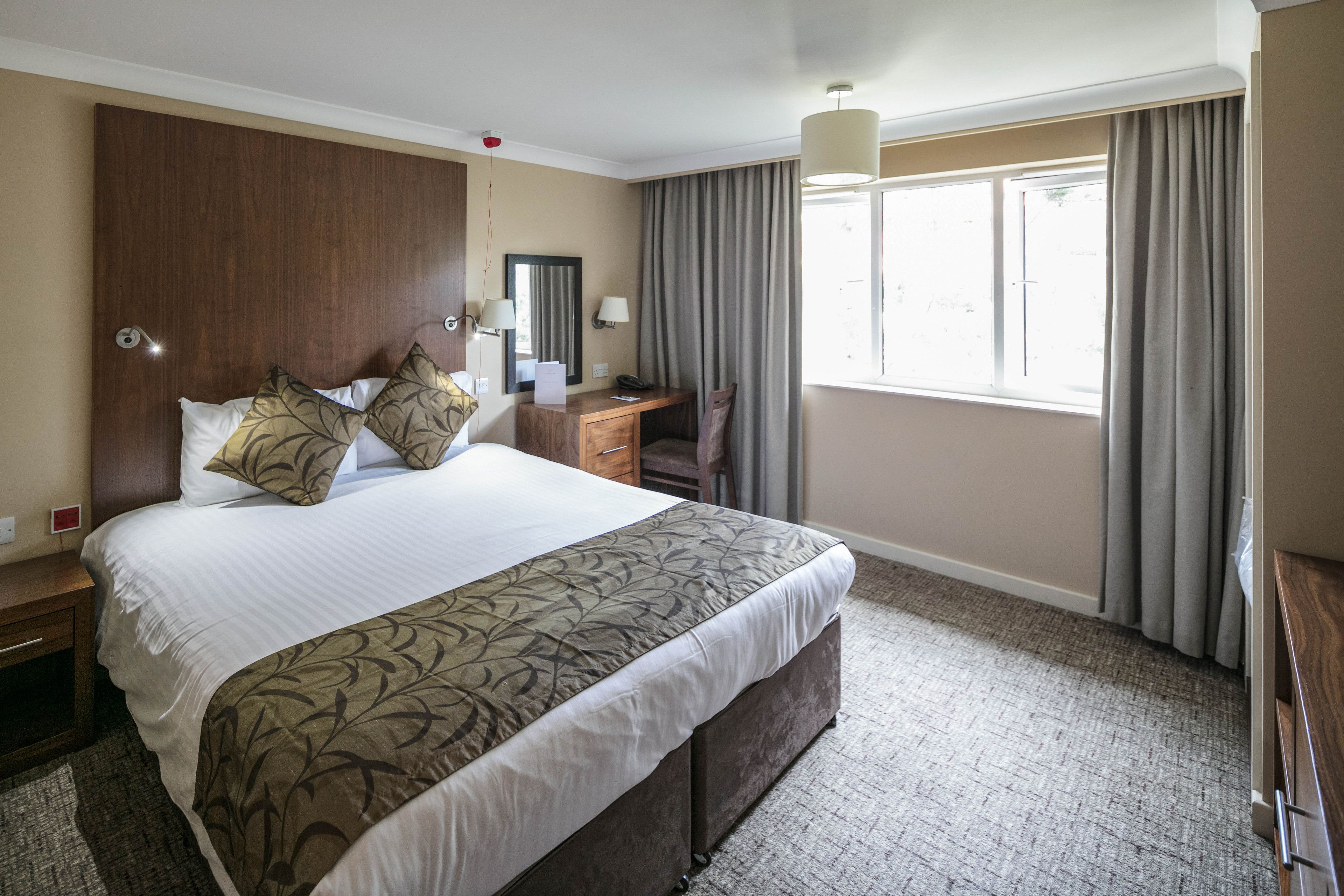 A hotel room with a large bed, a desk, a chair, a mirror, and a window with curtains.