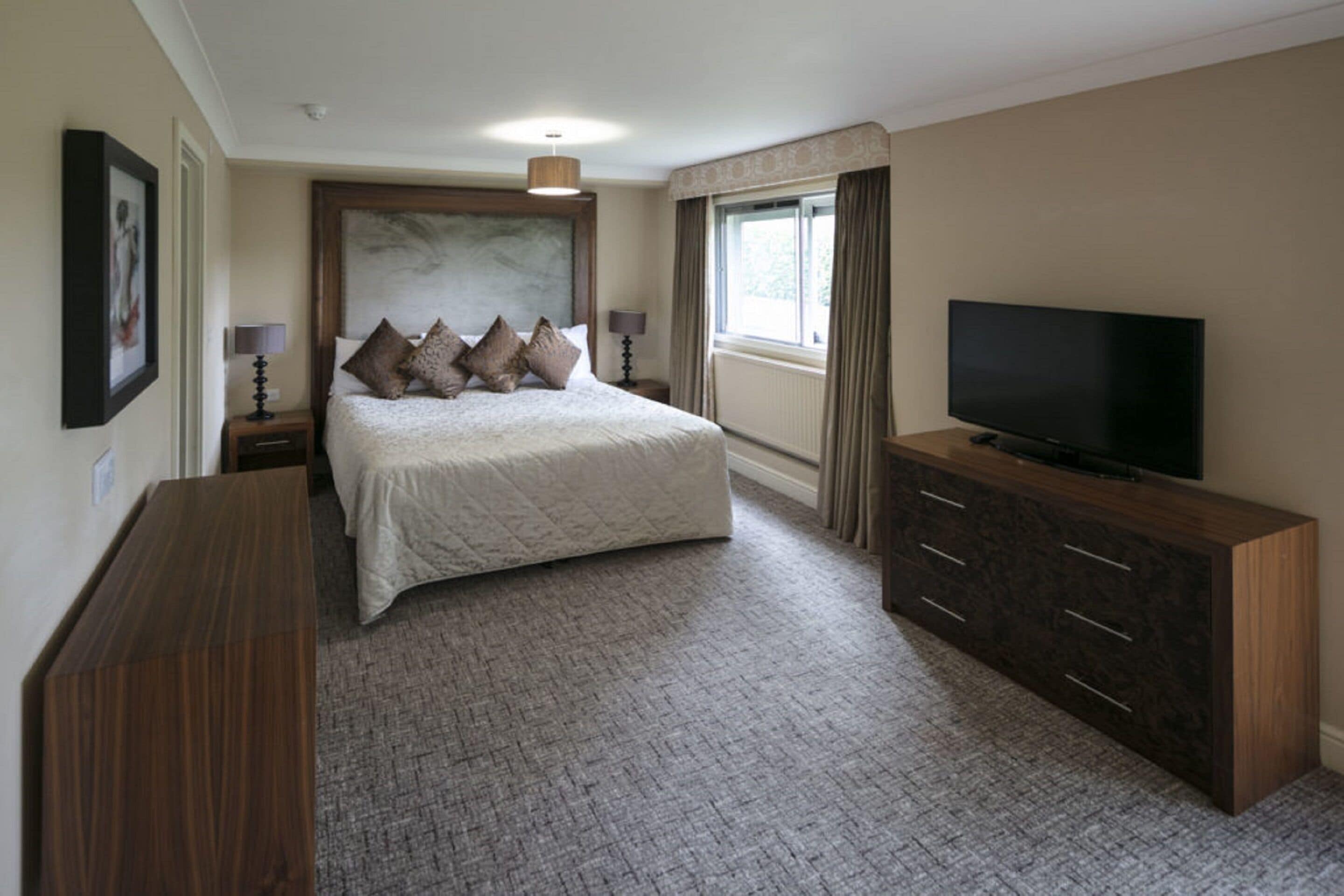 A hotel room with a large bed, a flat-screen TV, a wooden dresser, and a window with curtains.
