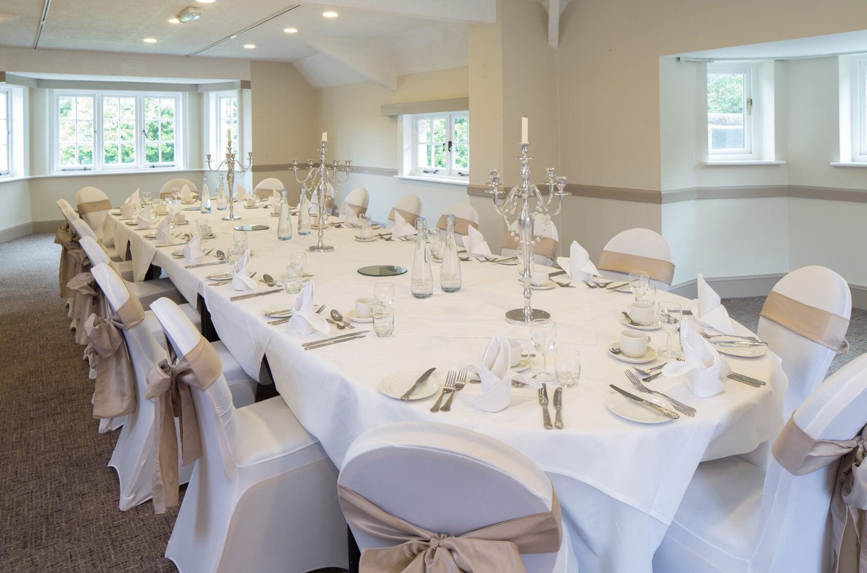 Une salle de banquet avec de longues tables dressées pour un événement formel, agrémentée de nappes blanches, de couverts en argent et de chandeliers.