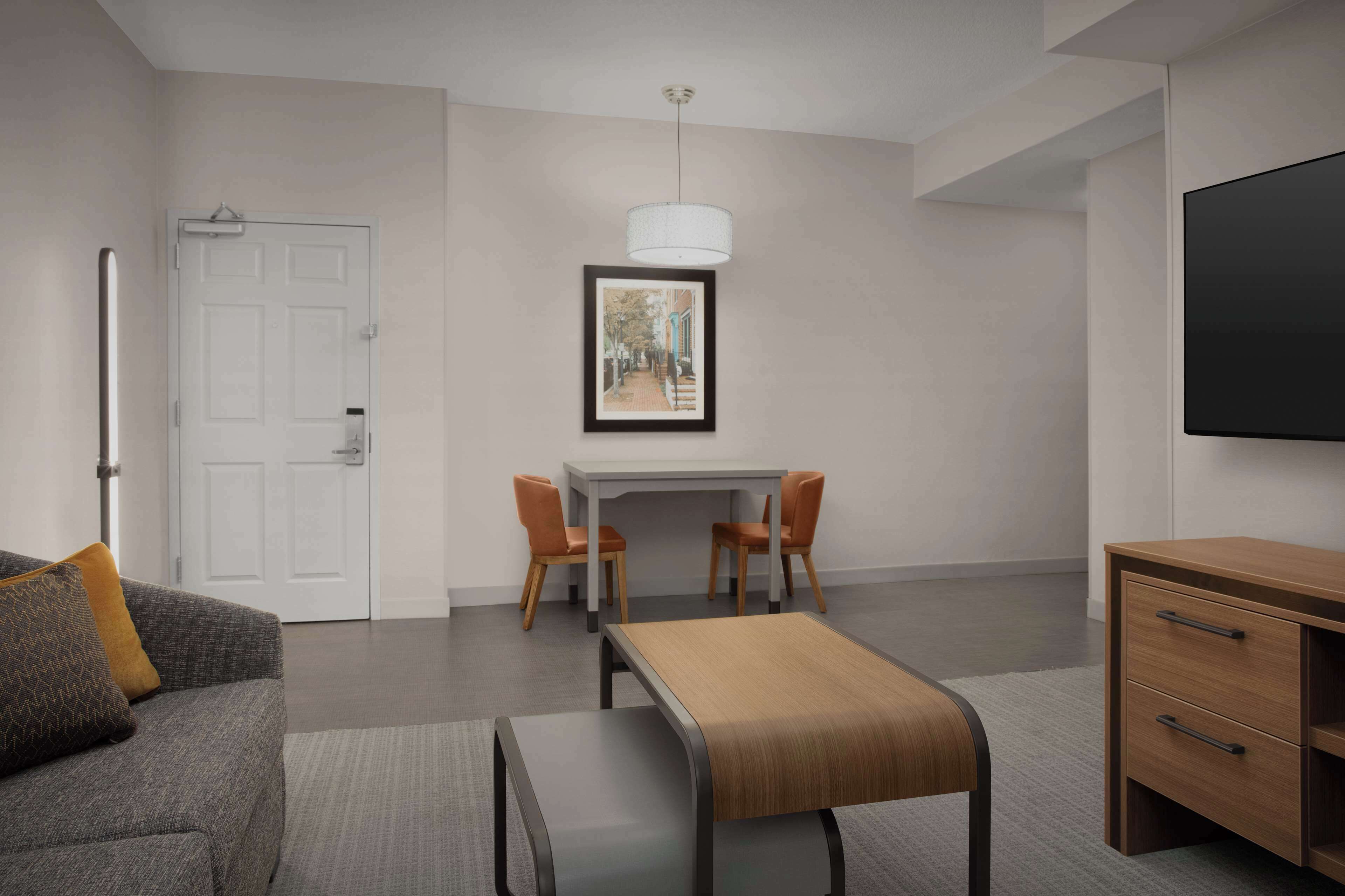 Une chambre d’hôtel moderne avec un canapé gris, une table basse en bois, un coin repas avec deux chaises, un tableau encadré au mur et un téléviseur sur le côté droit.