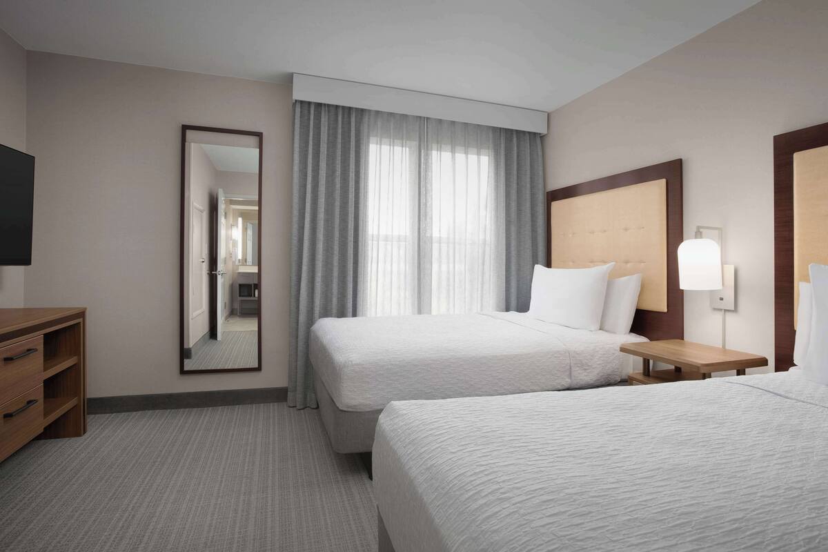 Una habitación de hotel con dos camas, un aparador de madera, un espejo y una ventana con cortinas.