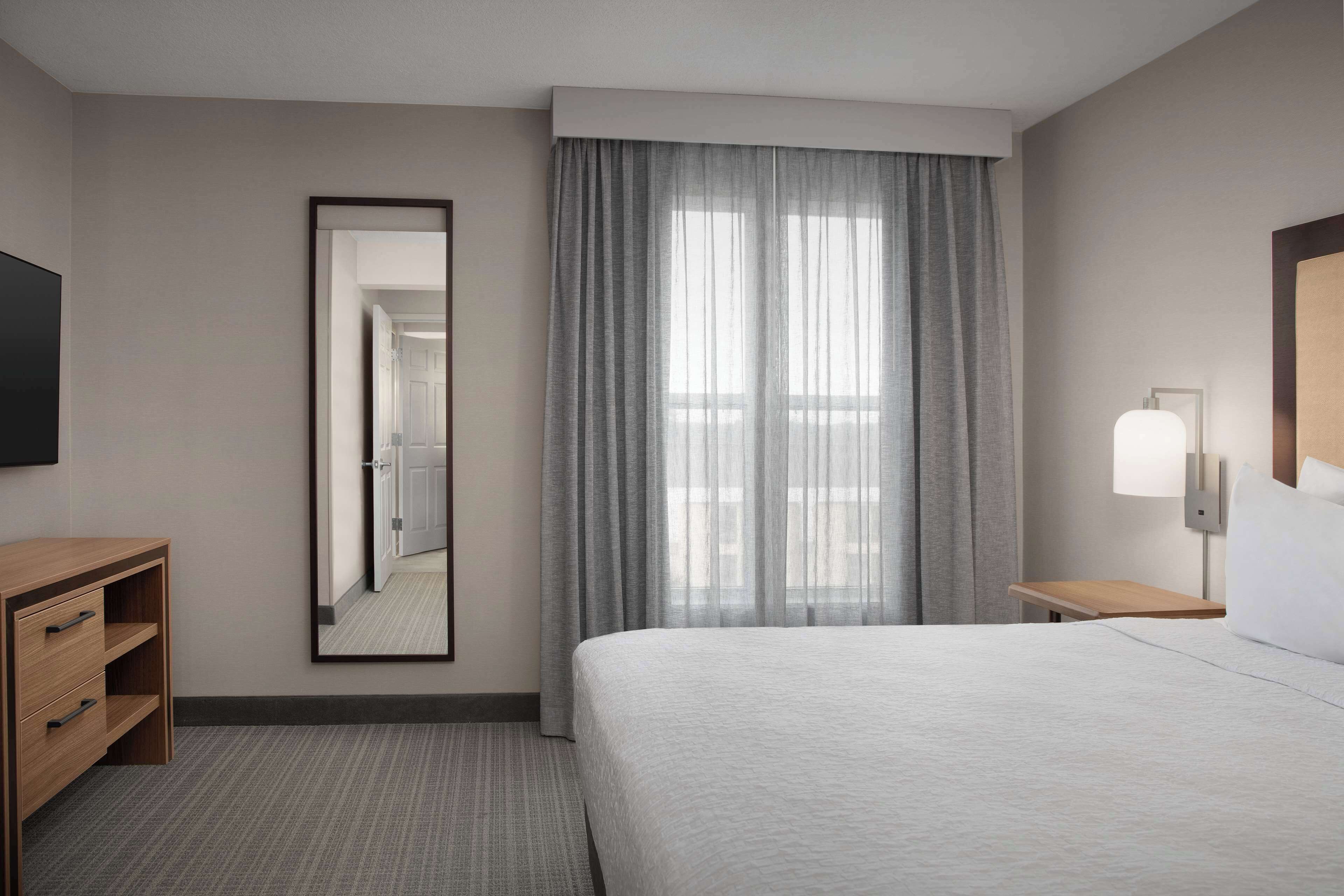 A hotel room with a large bed, a wooden nightstand with a lamp, a mirror, and a TV on the wall.