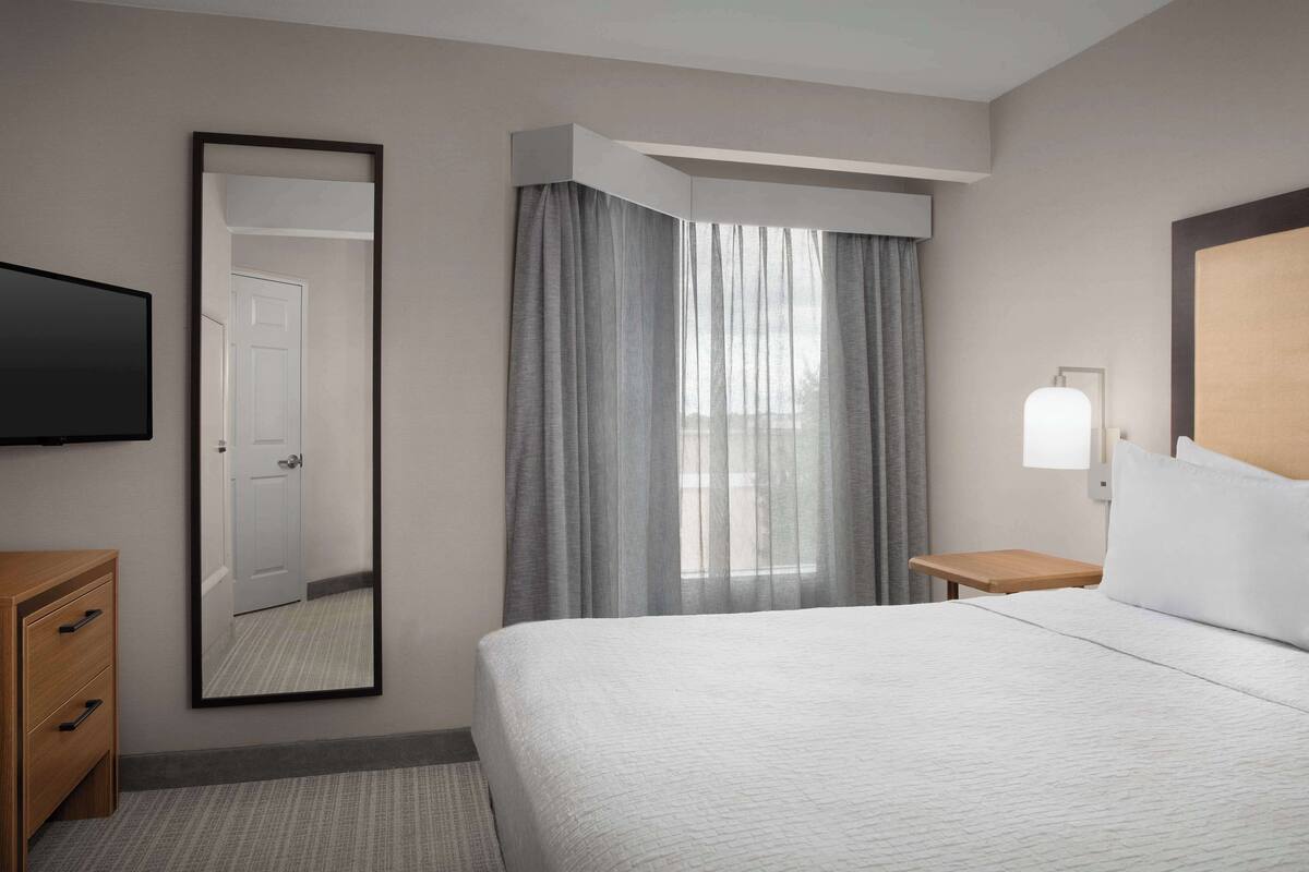 Habitación de hotel con cama, televisión, espejo, mesita de noche y ventana con cortinas.