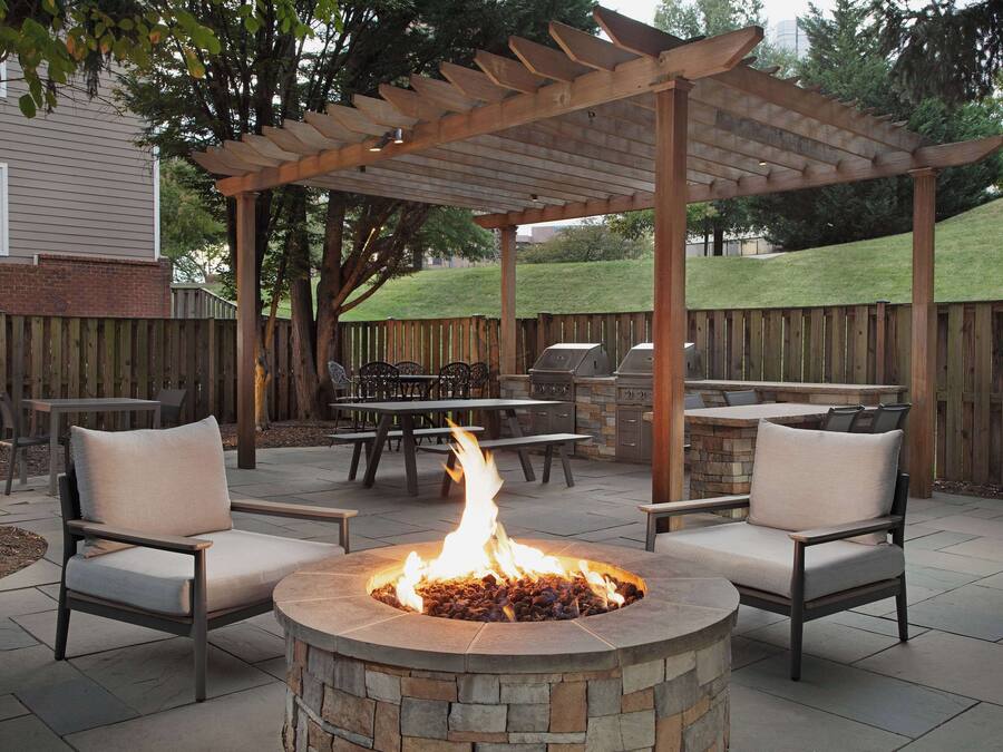 An outdoor patio with a stone fire pit, two chairs, a wooden pergola, and a barbecue grill.