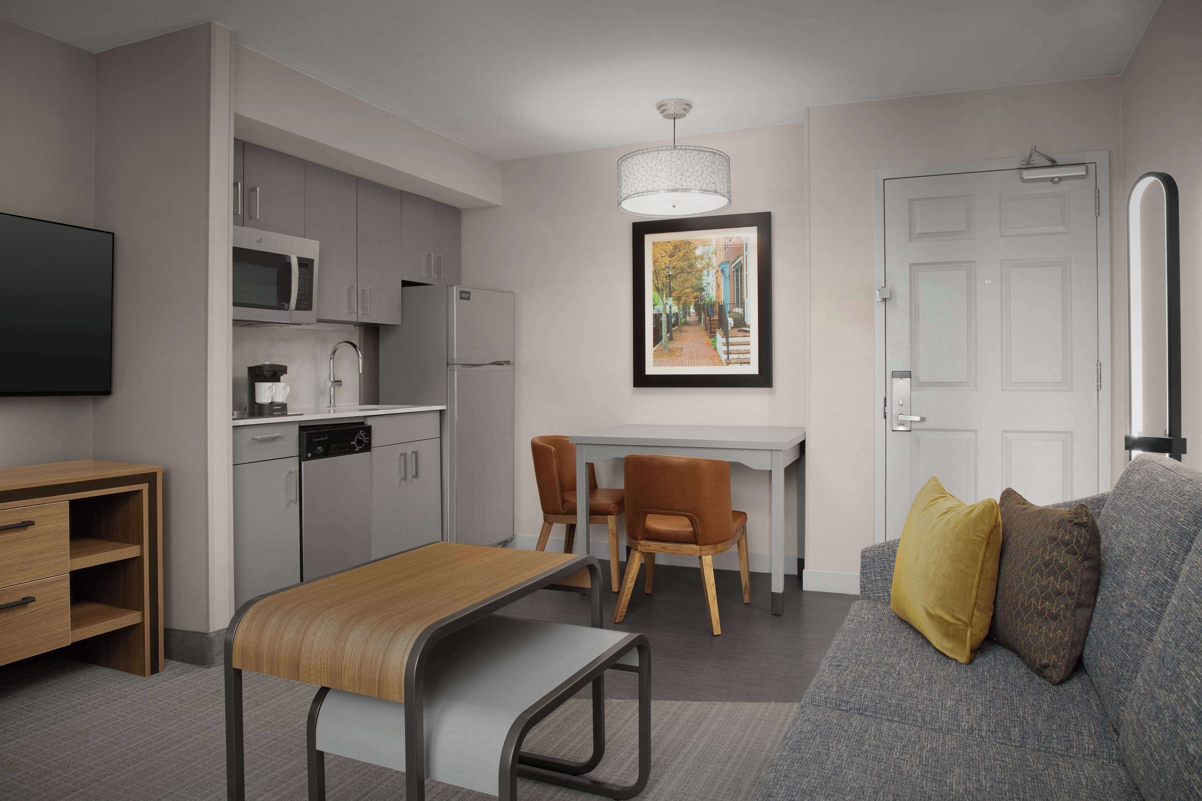 Une cuisine moderne avec des meubles gris, un four à micro-ondes et un lave-vaisselle. On y trouve une table à manger avec des chaises et un canapé avec des coussins. Un tableau encadré est accroché au mur, au-dessus de la table à manger.