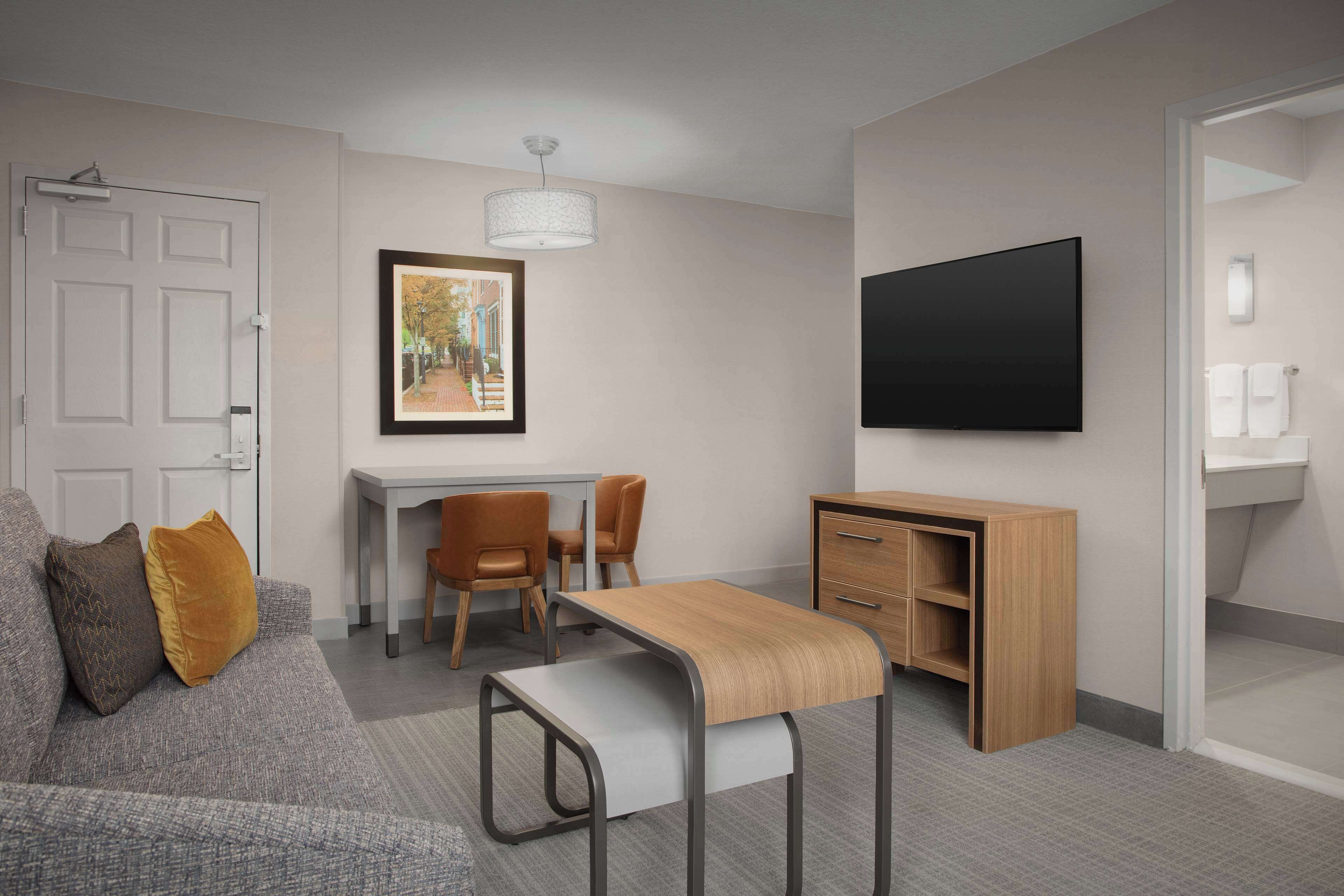 Una habitación de hotel moderna con un sofá gris, un escritorio pequeño con sillas, un televisor y un armario de madera.