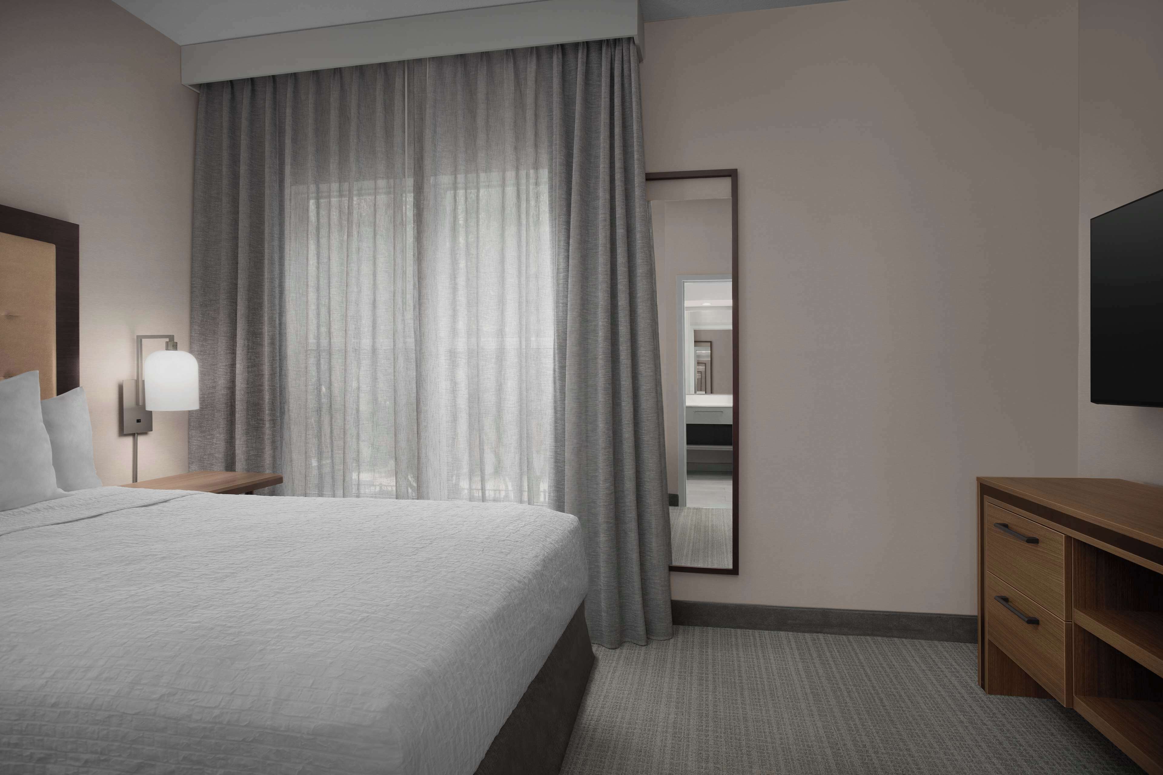 Une chambre d’hôtel avec un grand lit, une table de chevet avec une lampe, une commode en bois et un grand miroir au mur.