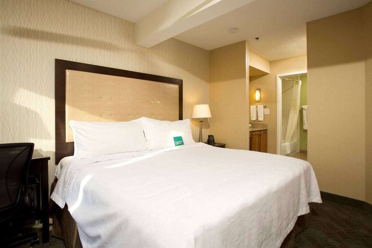 Habitación de hotel con cama, escritorio, silla, lámpara y baño visible a través de una puerta abierta.
