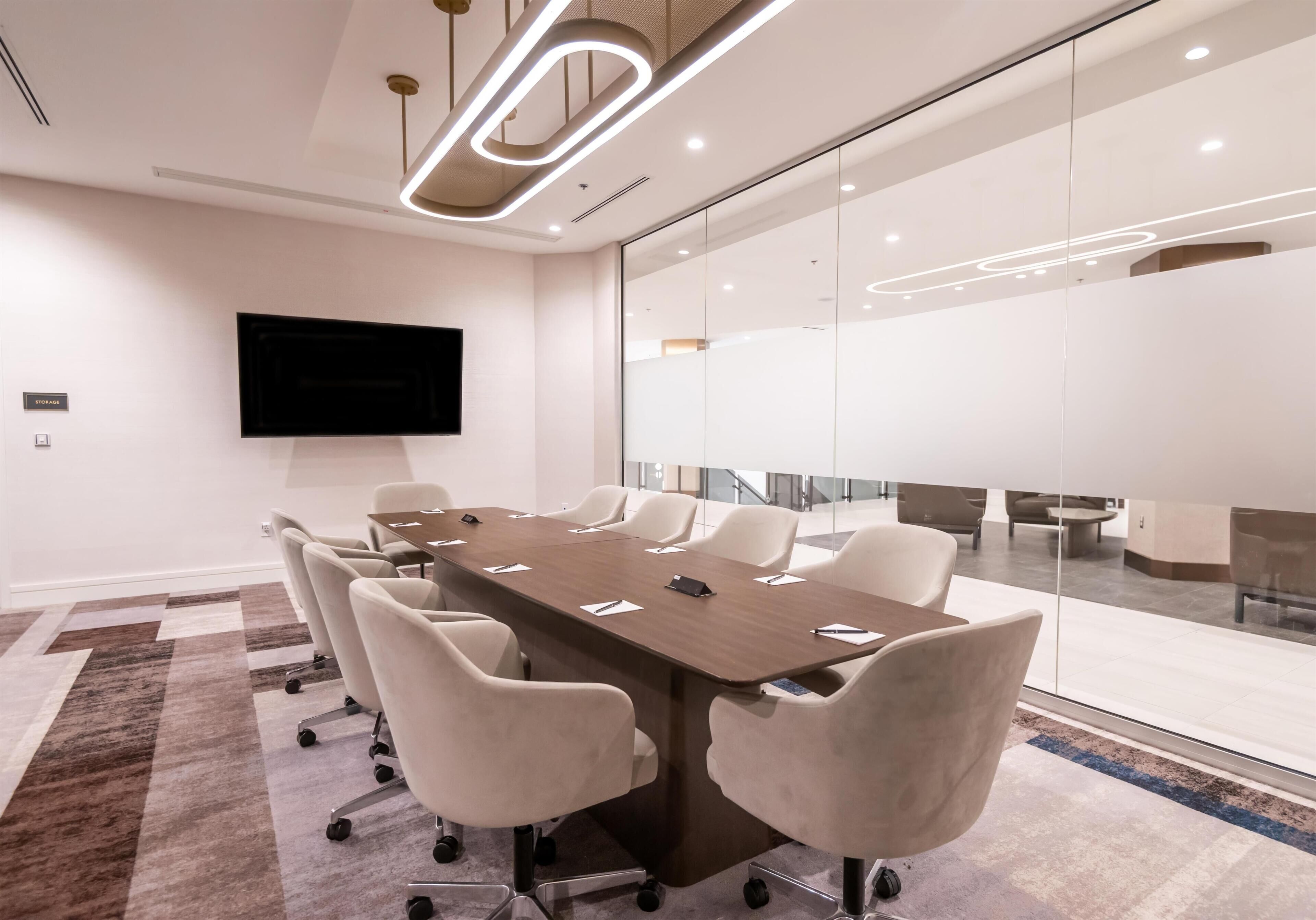 A conference room with a long table, white chairs, a flat-screen TV, and a glass wall.