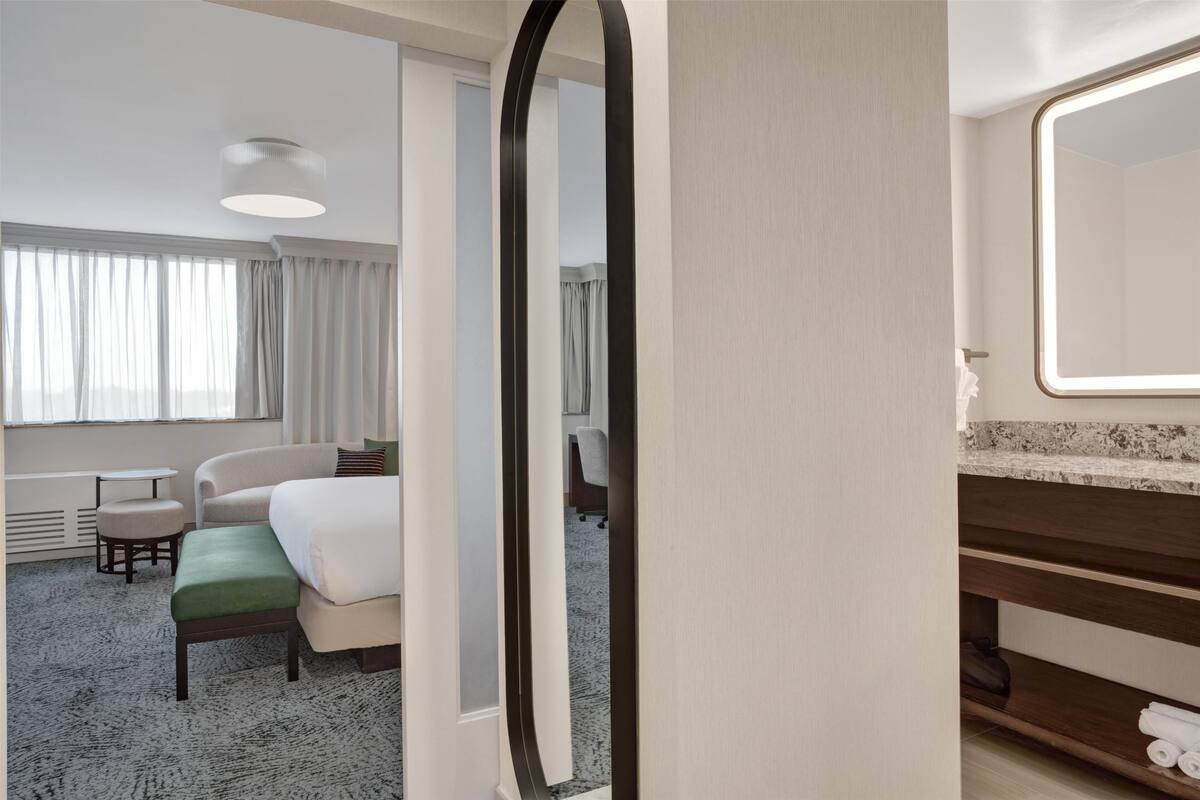 Habitaci ón de hotel con cama, una mesita, una silla, un espejo y baño con lavamanos y toallero.