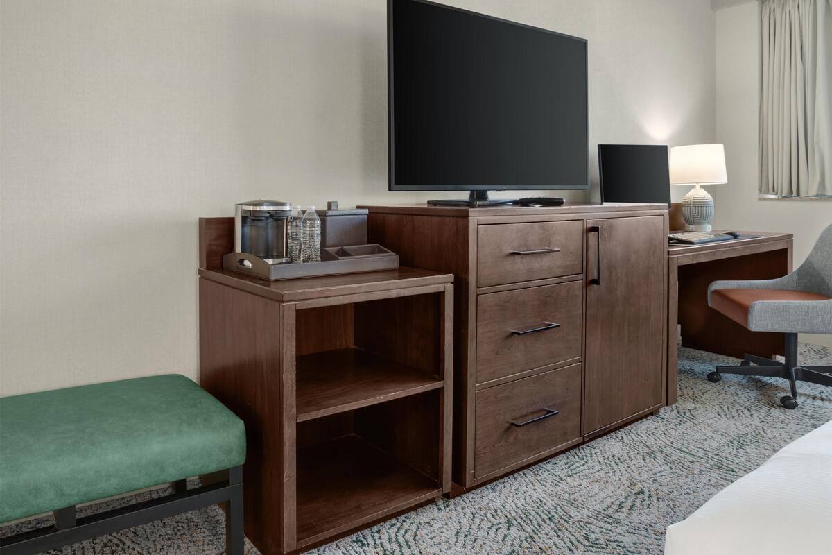 Habitación de hotel con escritorio de madera, televisor de pantalla plana, lámpara, banca verde y minibar.