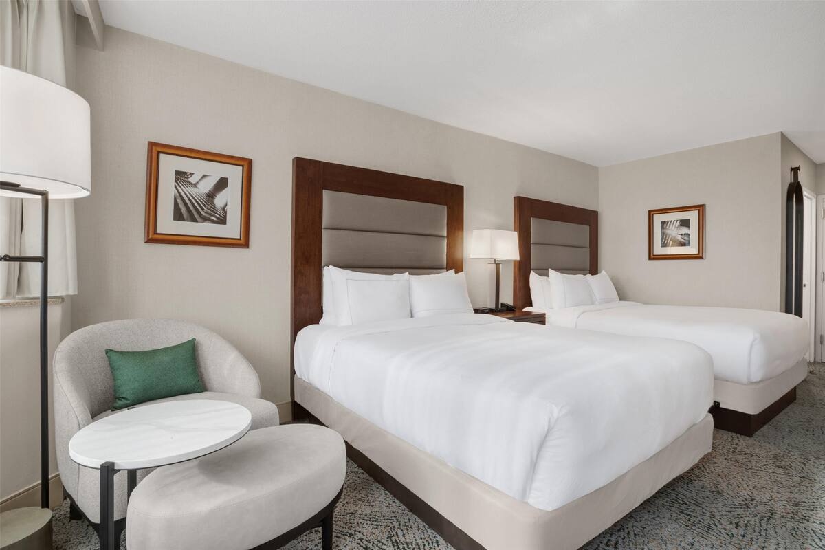 Habitación de hotel con dos camas, una mesita redonda pequeña, una silla y dos lámparas de noche.